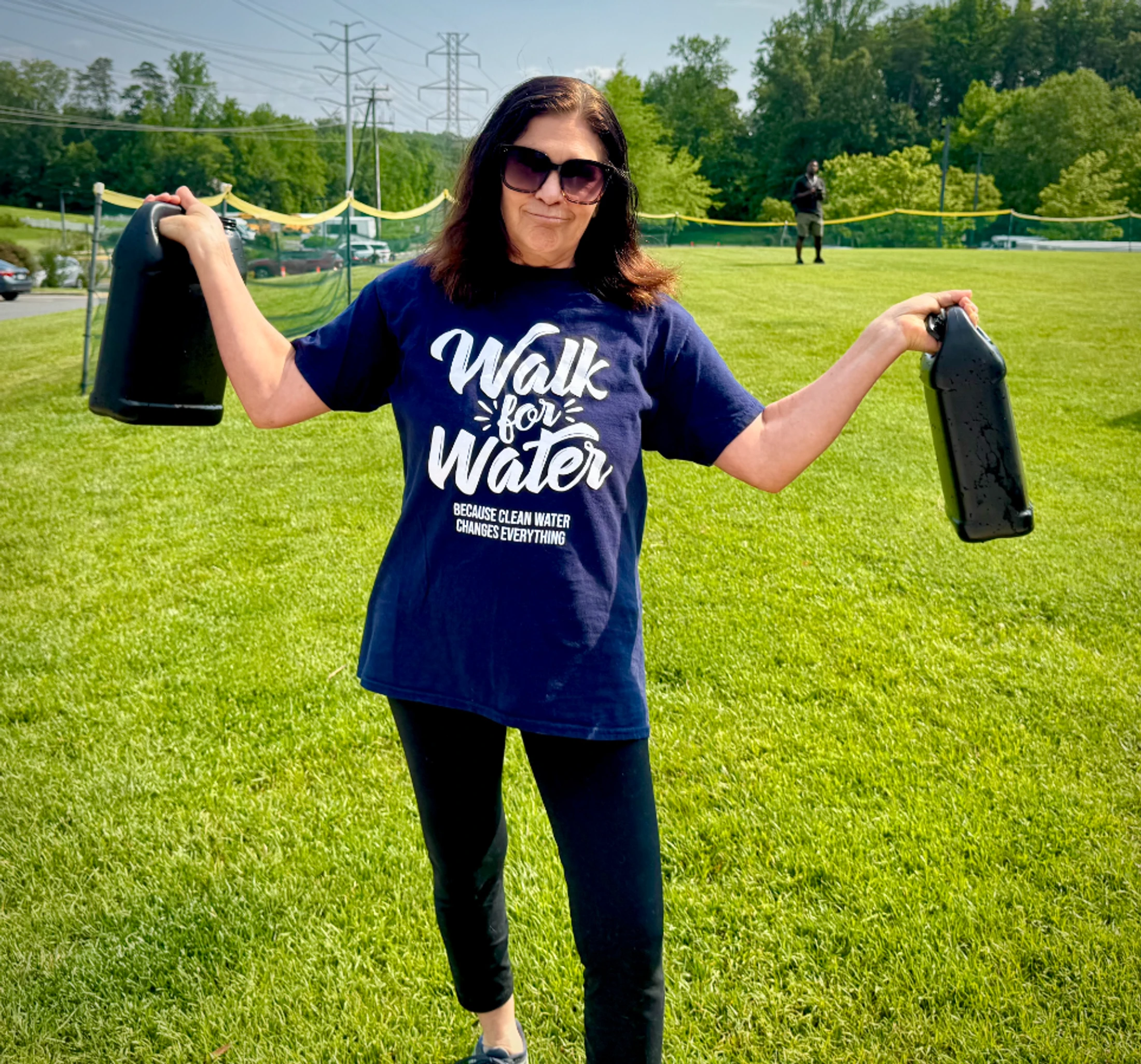 A woman in a blue 'Walk for Water' t-shirt and sunglasses holds two large black water jugs while standing in a grassy field on a sunny day.