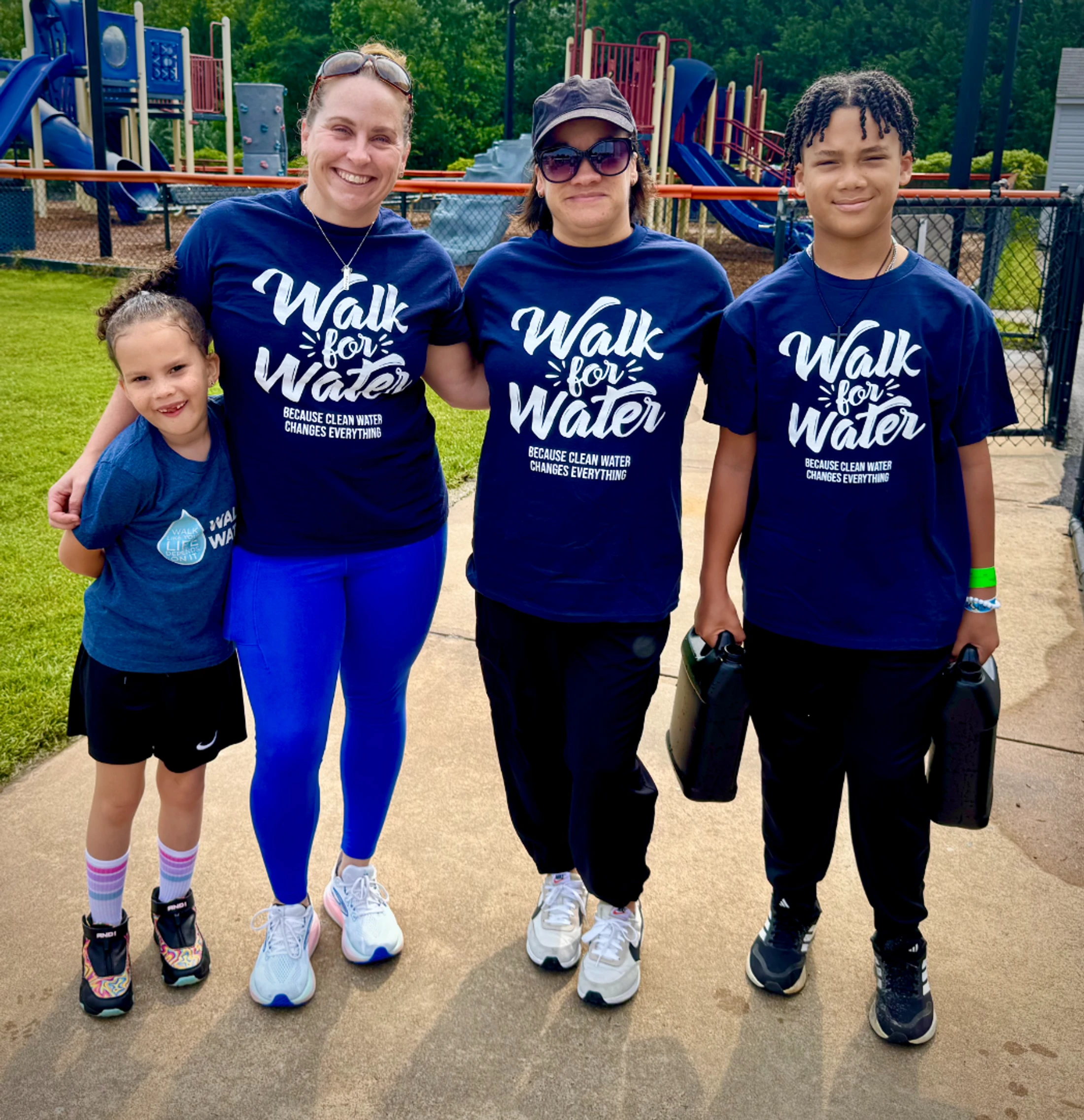 A group of two women and two children in matching 'Walk for Water' t-shirts pose for a photo on a path, with a playground in the background.
