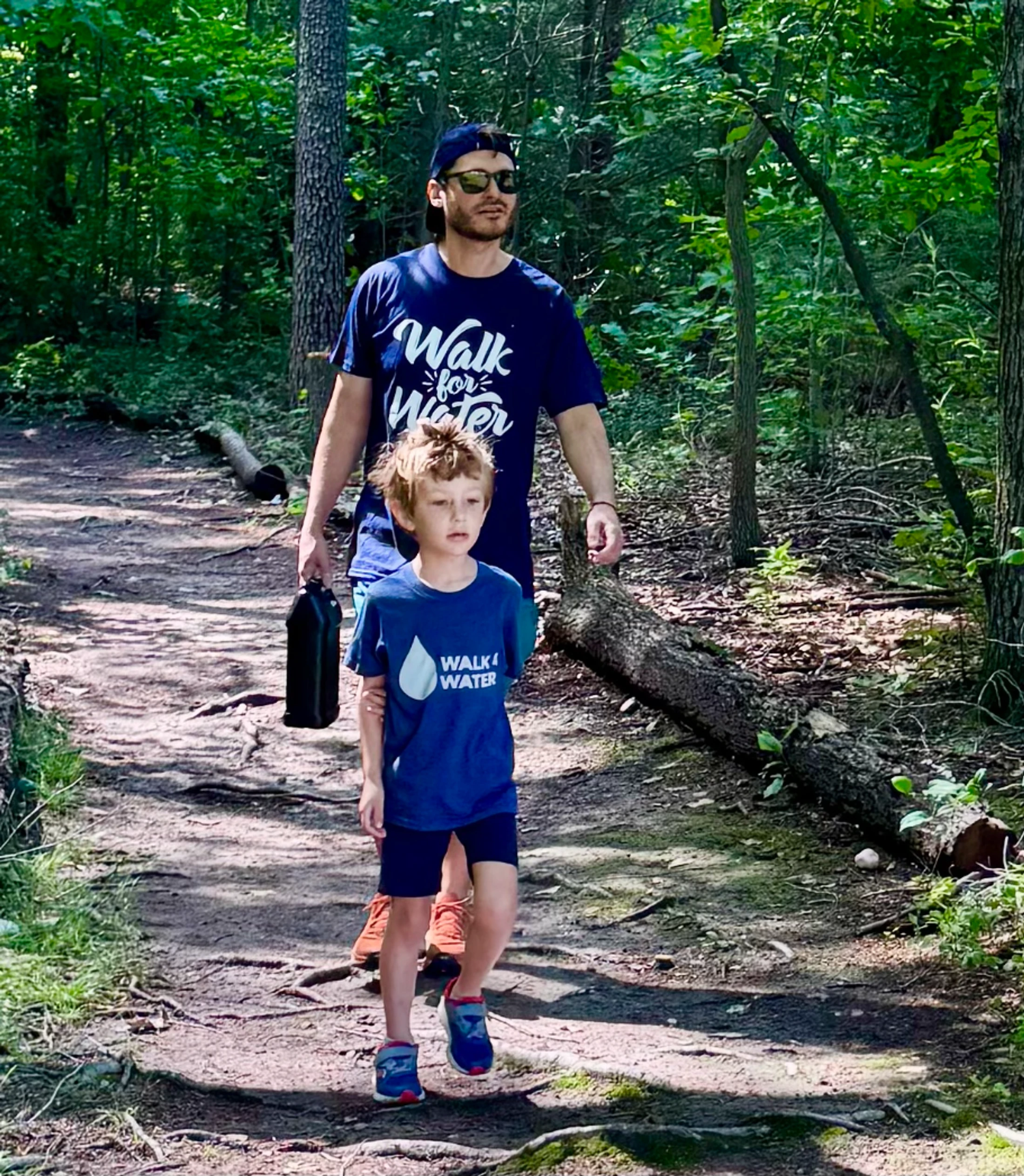 A man and a young boy in matching blue 'Walk for Water' shirts hike together on a dirt trail through a sunlit forest.
