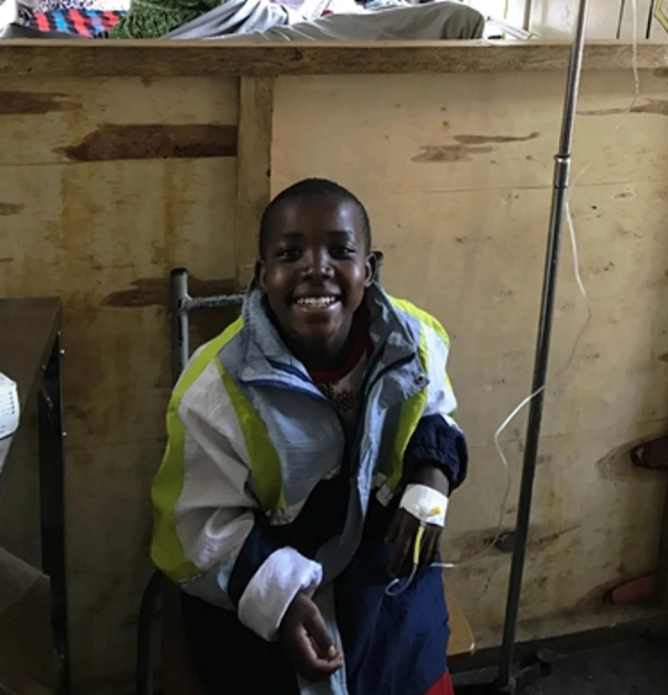 A young Black boy in a jacket smiles widely while sitting in a chair. He has an intravenous drip attached to his hand in what looks like a simple clinic.