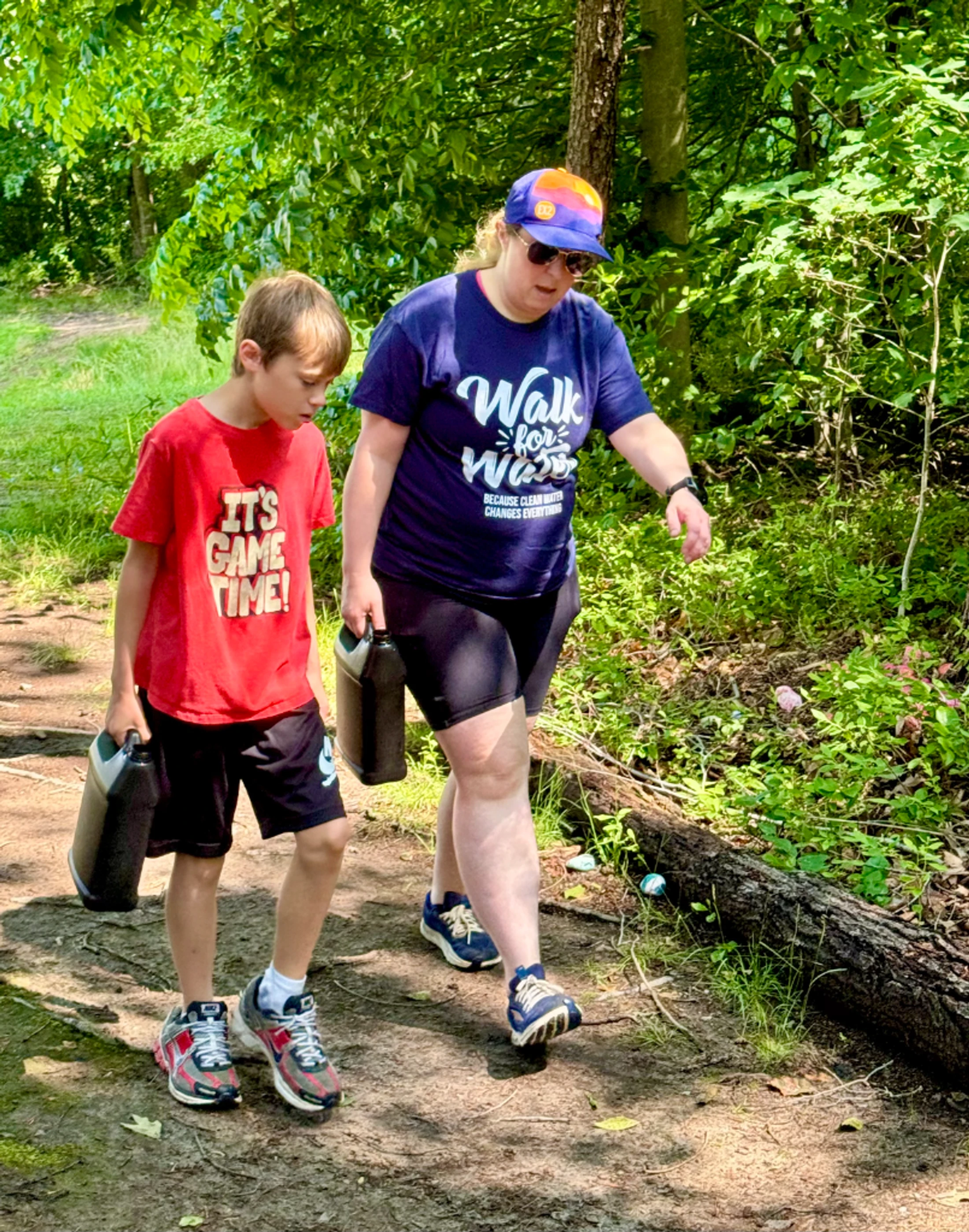 A woman and a young boy participate in a 'Walk for Water' event, carrying black jugs as they walk along a dirt path in a sunny, wooded area.
