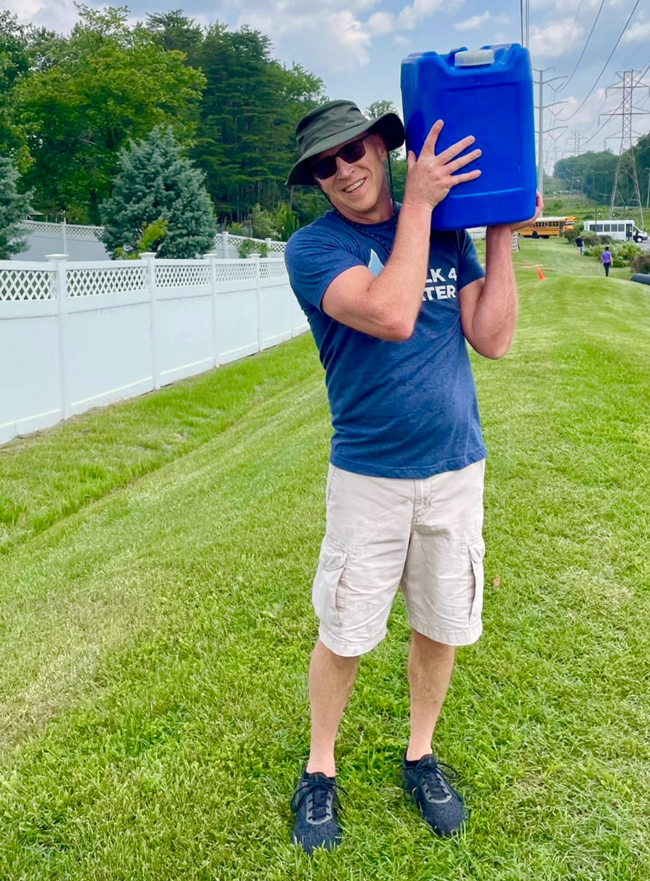 A smiling man wearing a hat and sunglasses carries a large blue water container on his shoulder while walking on a grassy lawn for a charity event.