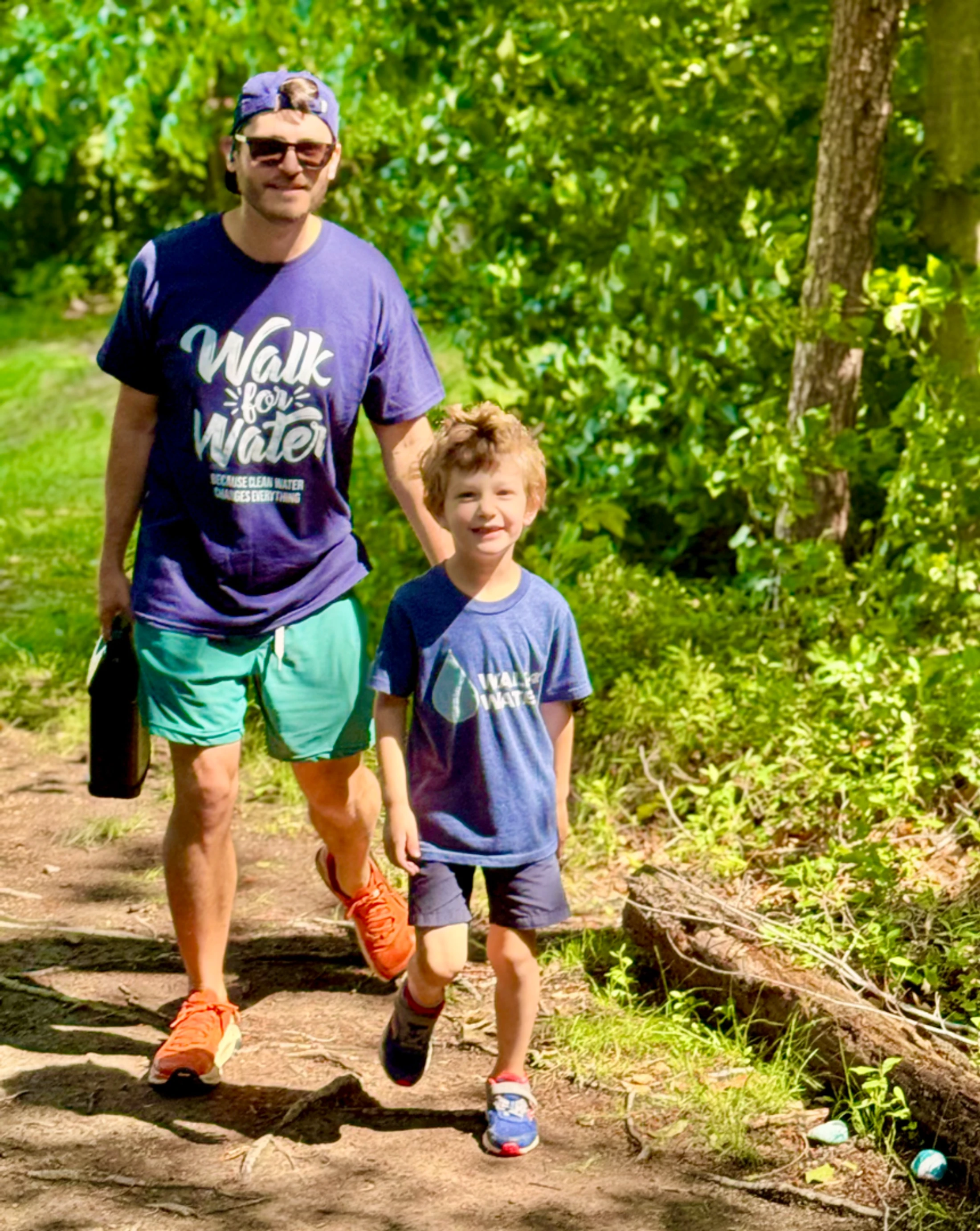 A father and his young son walk together on a dirt trail through a sunny, green forest. They are both wearing matching 'Walk for Water' t-shirts.