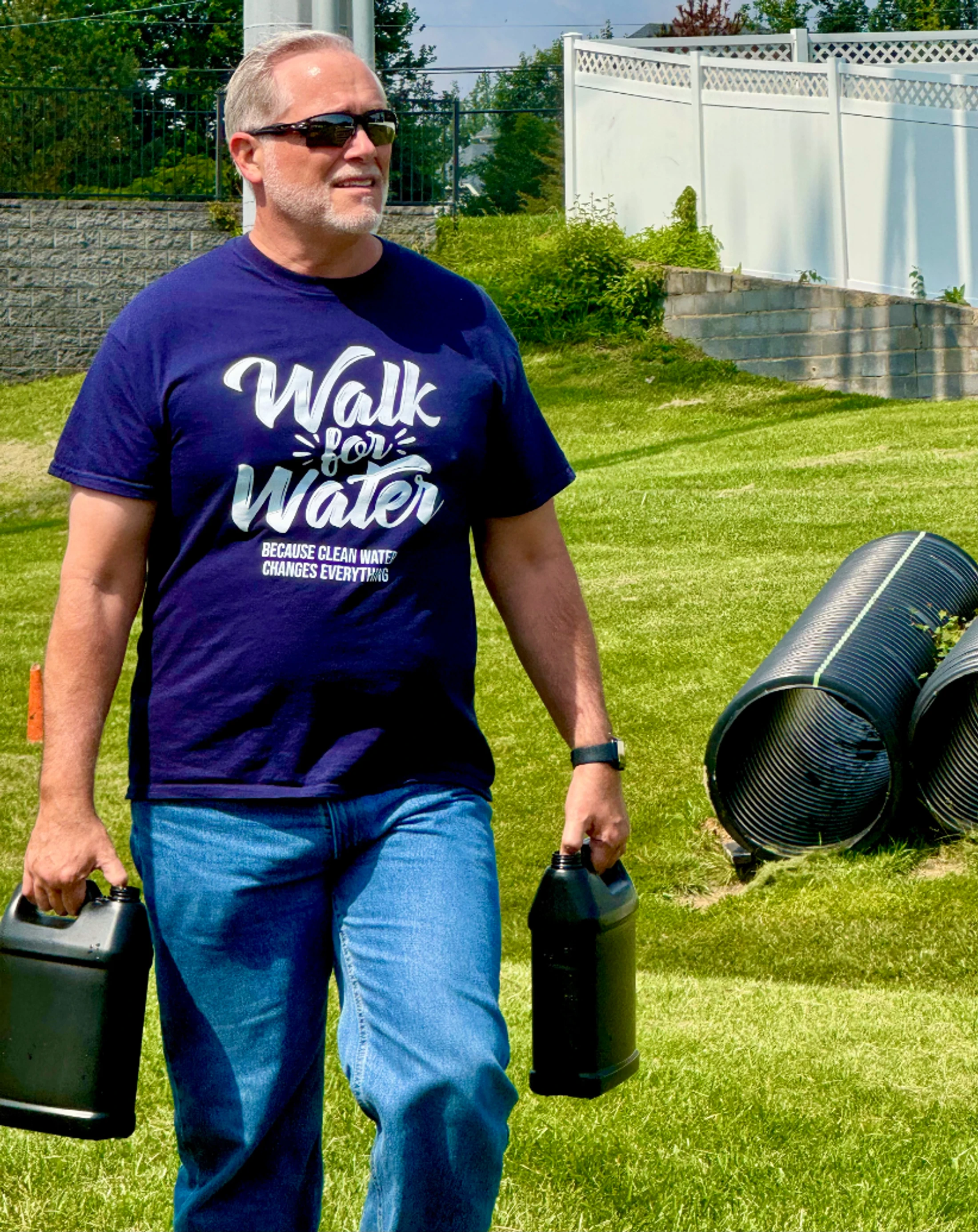 A man in sunglasses and a blue "Walk for Water" t-shirt walks across a grassy lawn carrying two large black plastic jugs, one in each hand.