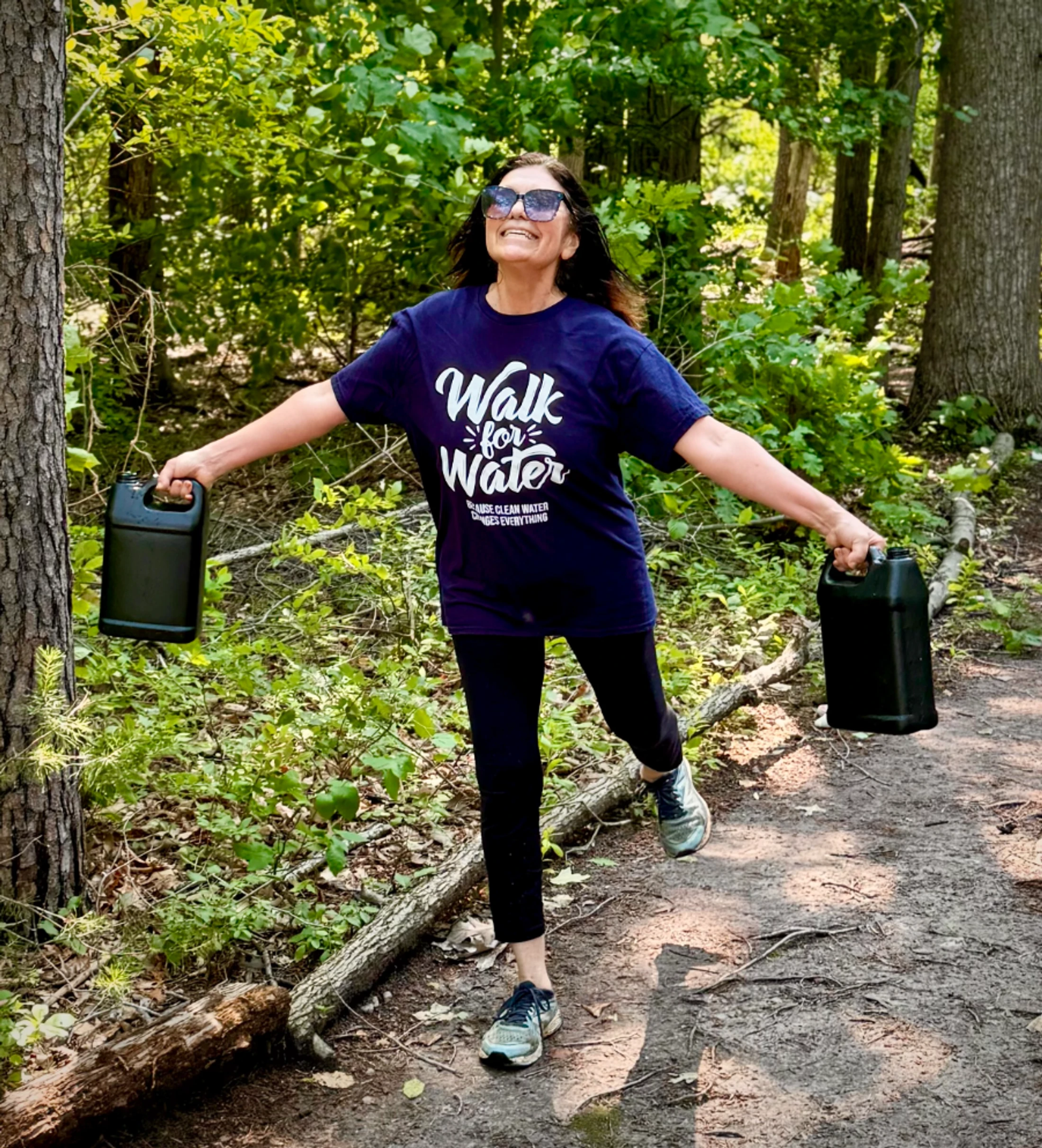 A smiling woman wearing a 'Walk for Water' t-shirt carries two large black water jugs while walking on a dirt path through a lush green forest.