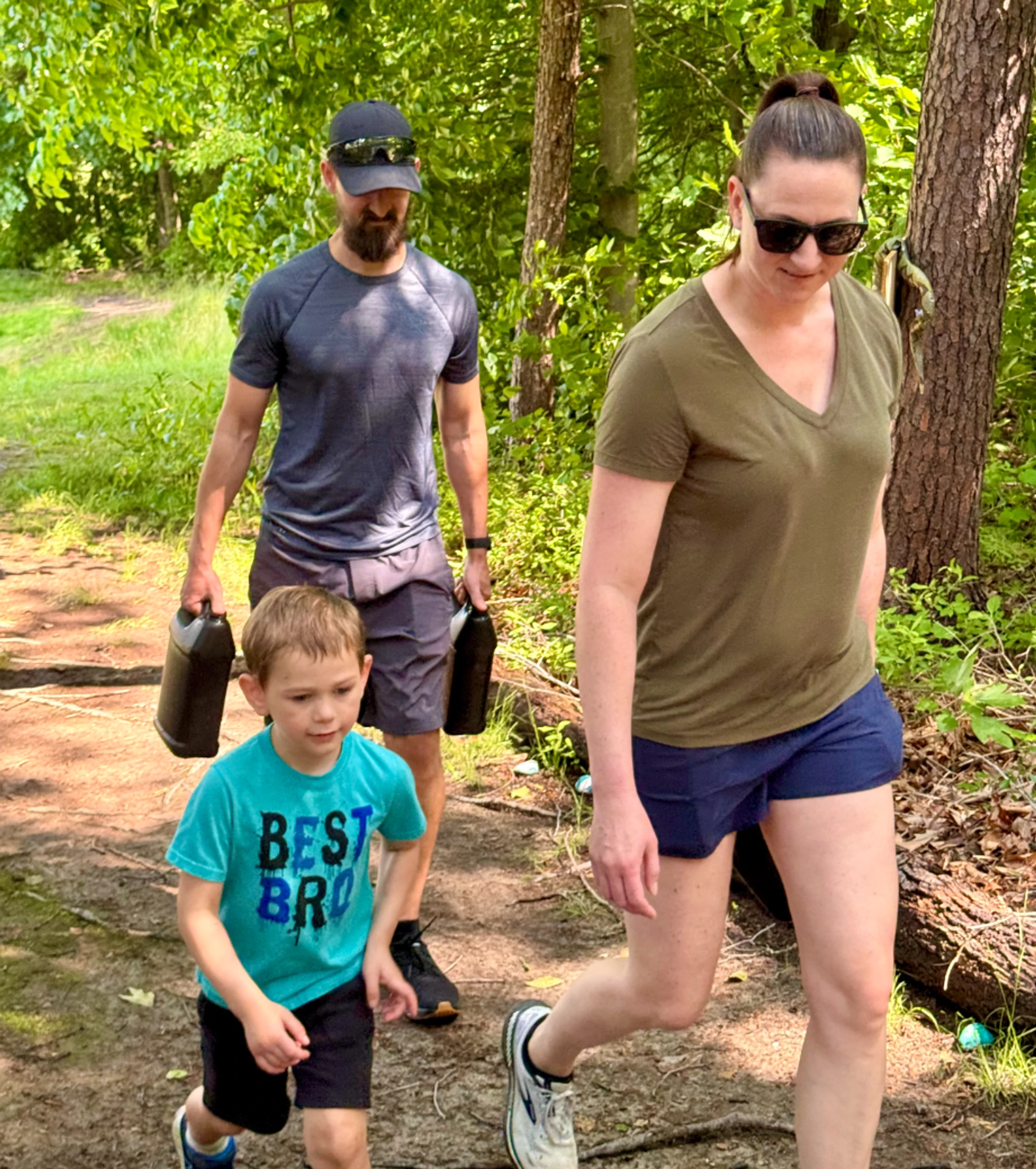 A family of three, a man, a woman, and a young boy, are hiking on a dirt path through a sunlit forest. The man is carrying two black jugs.