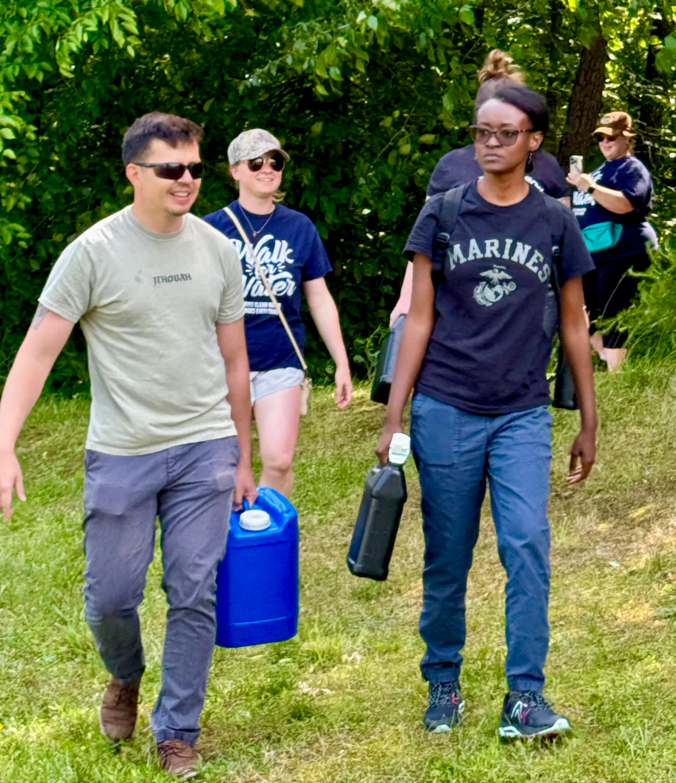 A group of people walk on a grassy path during an outdoor event, with a man and woman in front carrying large blue and black water jugs.