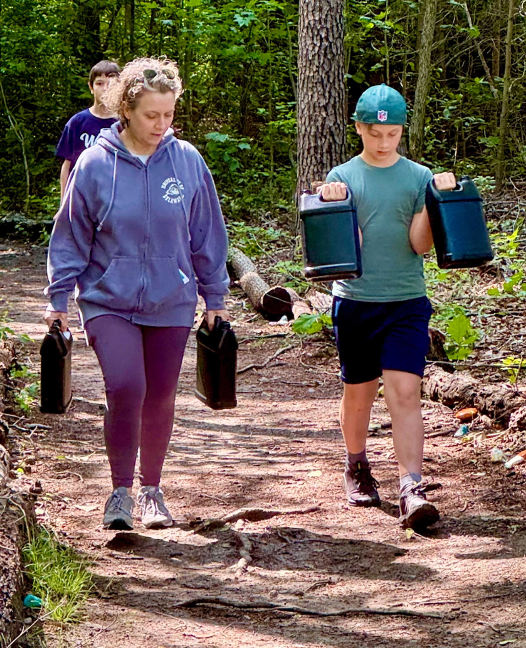 A woman and a young boy walk side-by-side on a dirt path in the woods, each carrying two heavy black jugs. Another boy walks behind them.