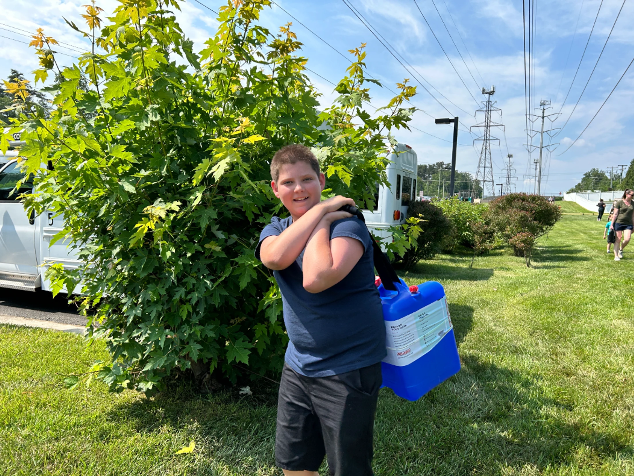 A smiling boy in a blue shirt stands on a grassy lawn next to a large bush, carrying a big blue plastic container over his shoulder on a sunny day.