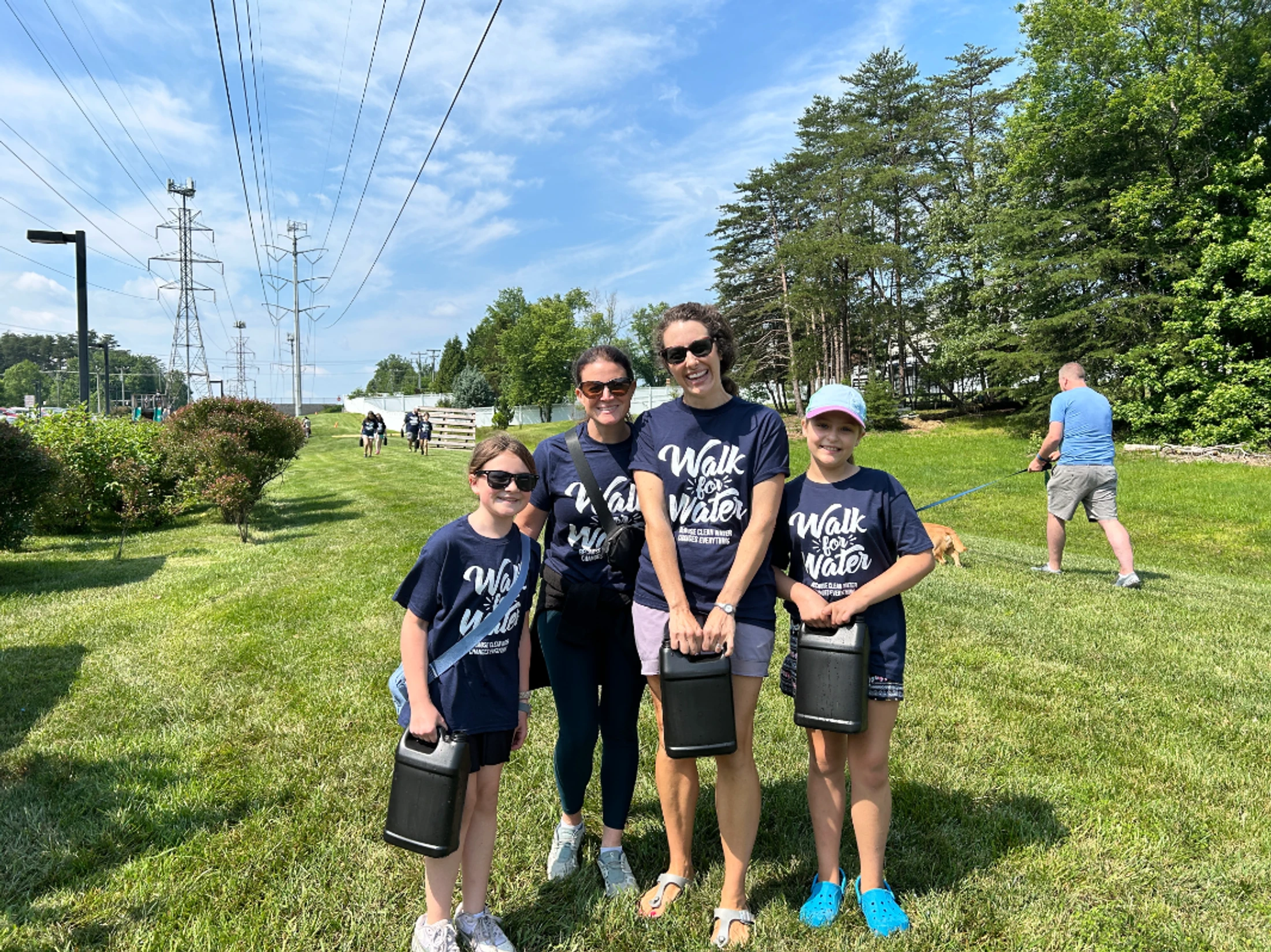 Two women and two girls in matching 'Walk for Water' t-shirts smile while holding water containers at an outdoor charity walk on a sunny day.
