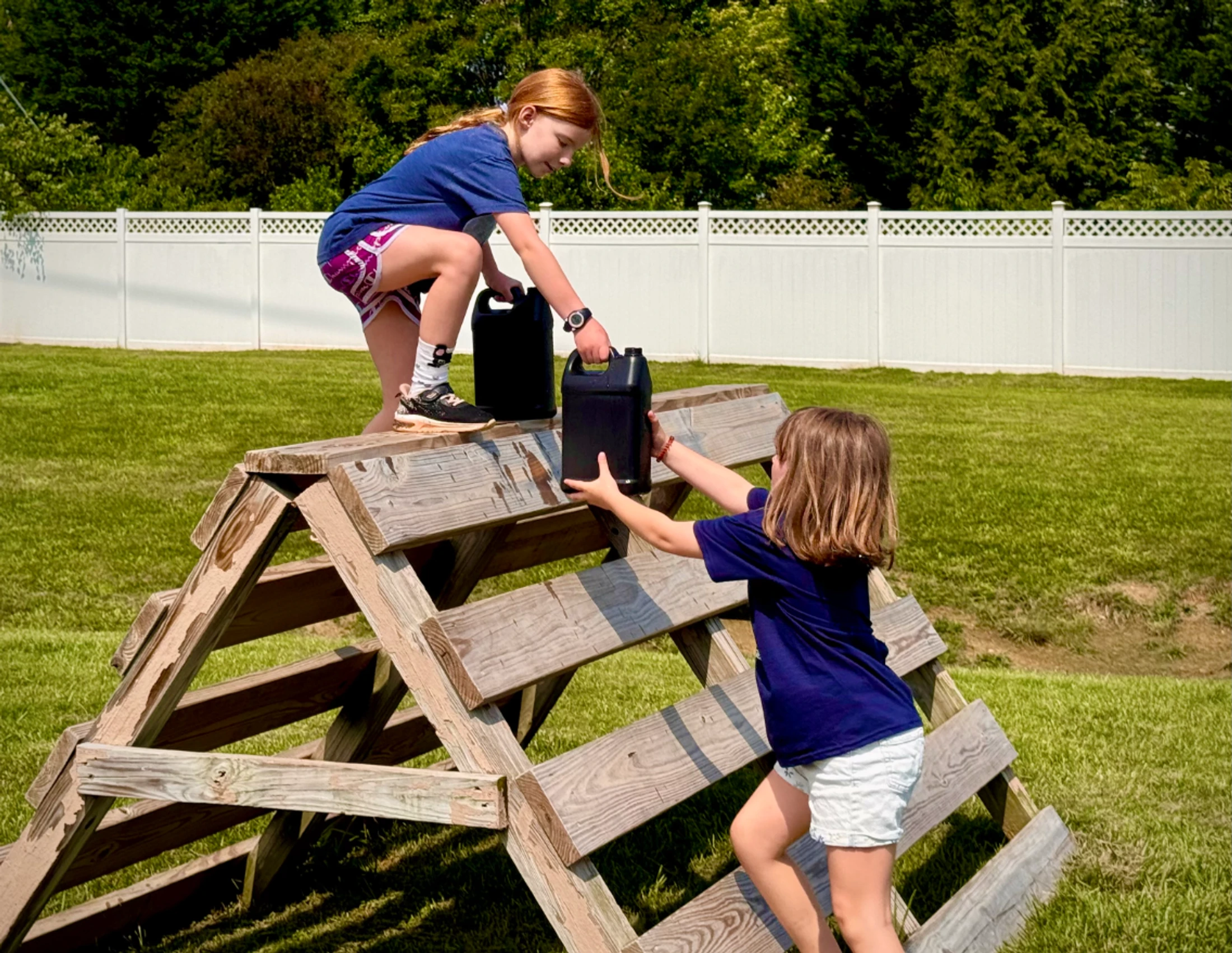 Two young girls play on a wooden A-frame structure in a grassy yard. One girl passes a black container to another girl who is on top of the structure.
