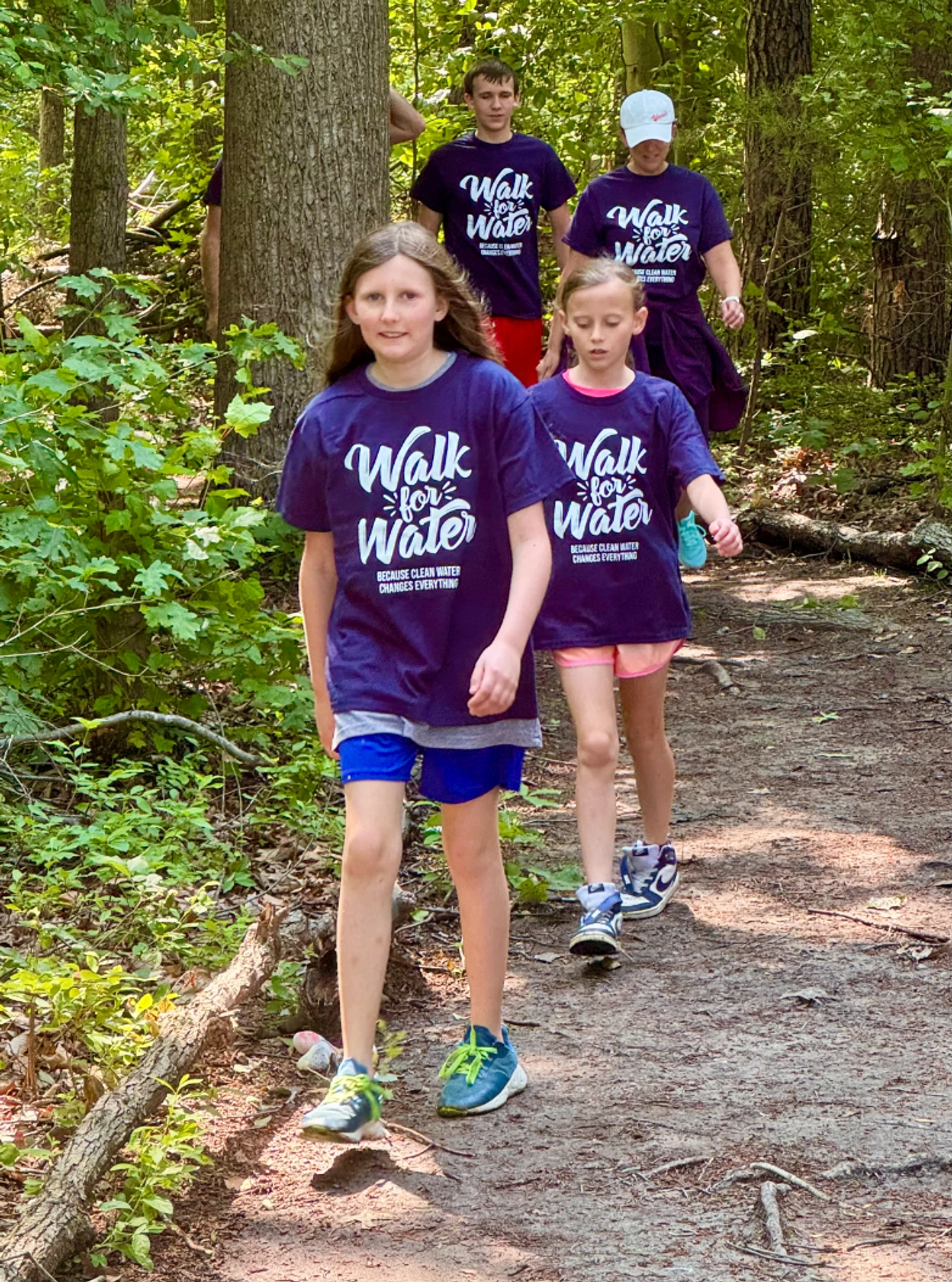 A group of adults and children in matching purple 'Walk for Water' t-shirts hike along a narrow dirt path through a sunlit forest.