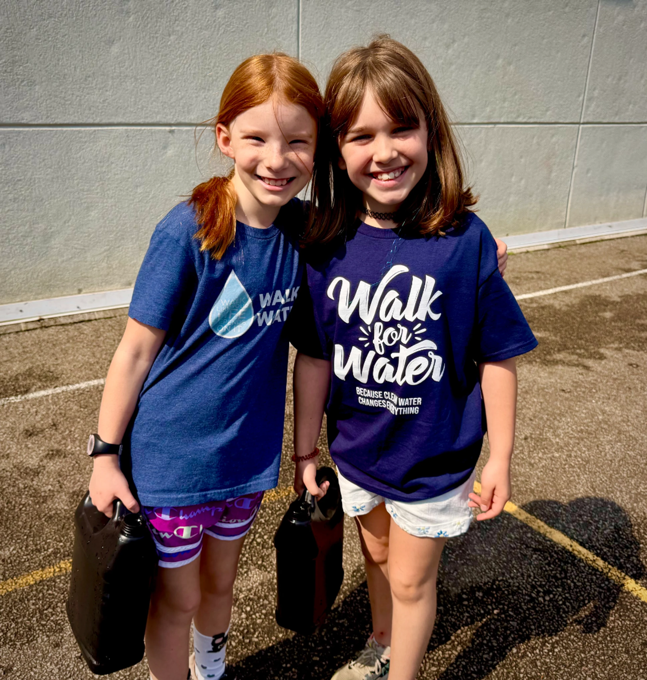 Two young girls smile at the camera while holding black water containers. They are wearing blue t-shirts for a 'Walk for Water' charity event.