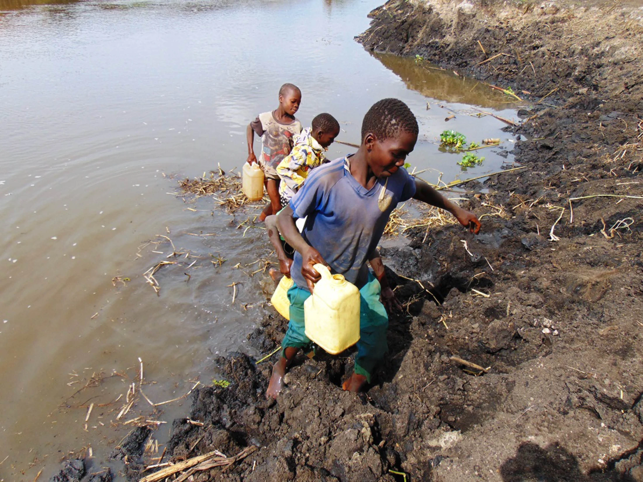 Three young children carry yellow jerrycans as they collect water from a muddy river, with one boy climbing up the slippery bank in the foreground.