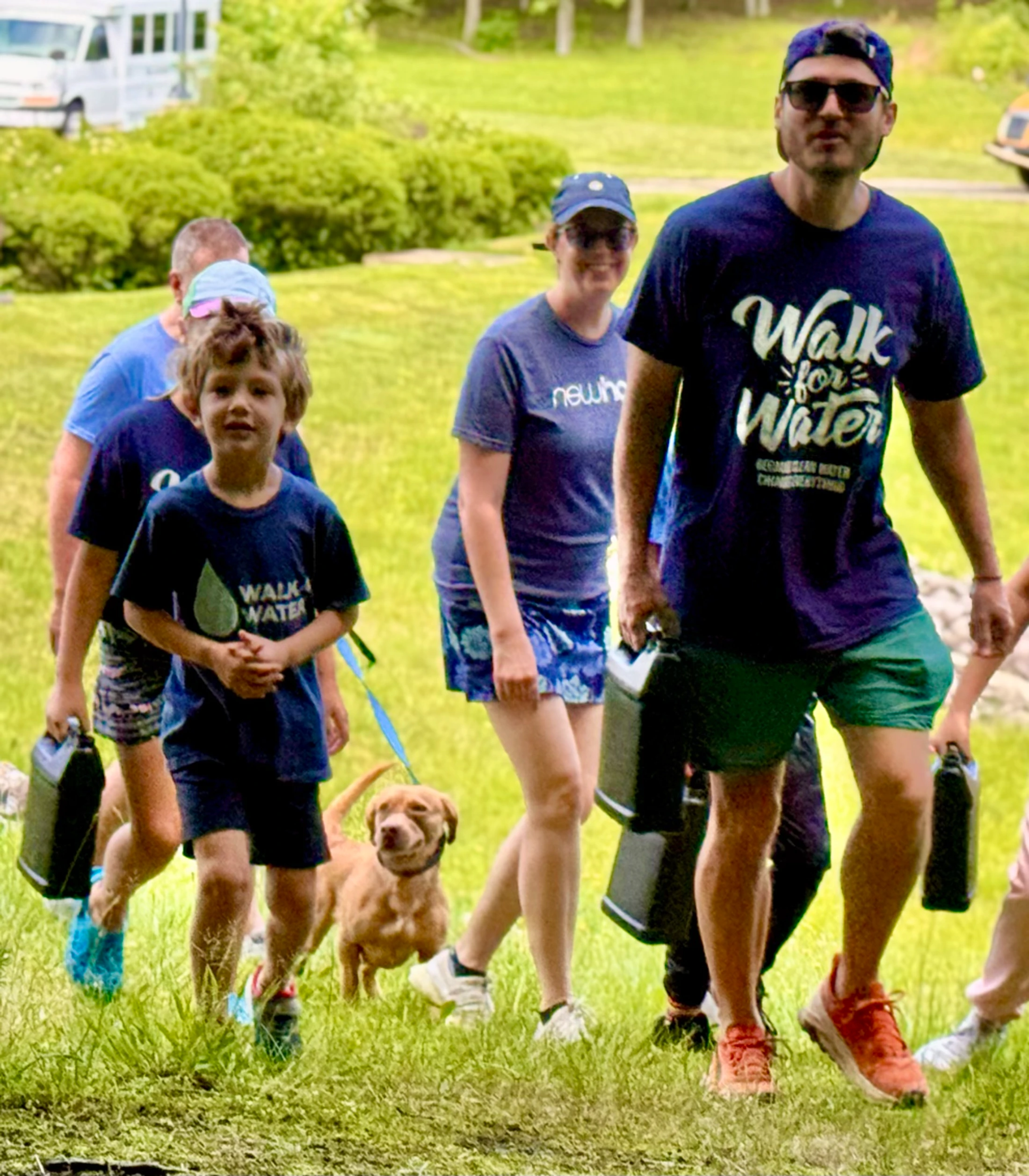 A group of adults, a child, and a small dog walk across a grassy field wearing matching 'Walk for Water' t-shirts and carrying black water containers.