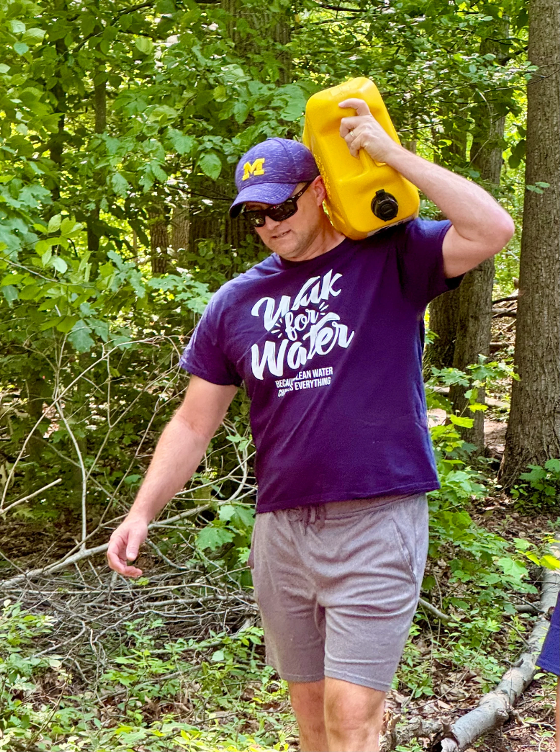 A man in a purple 'Walk for Water' t-shirt carries a large yellow water jug over his shoulder while walking through a wooded area.