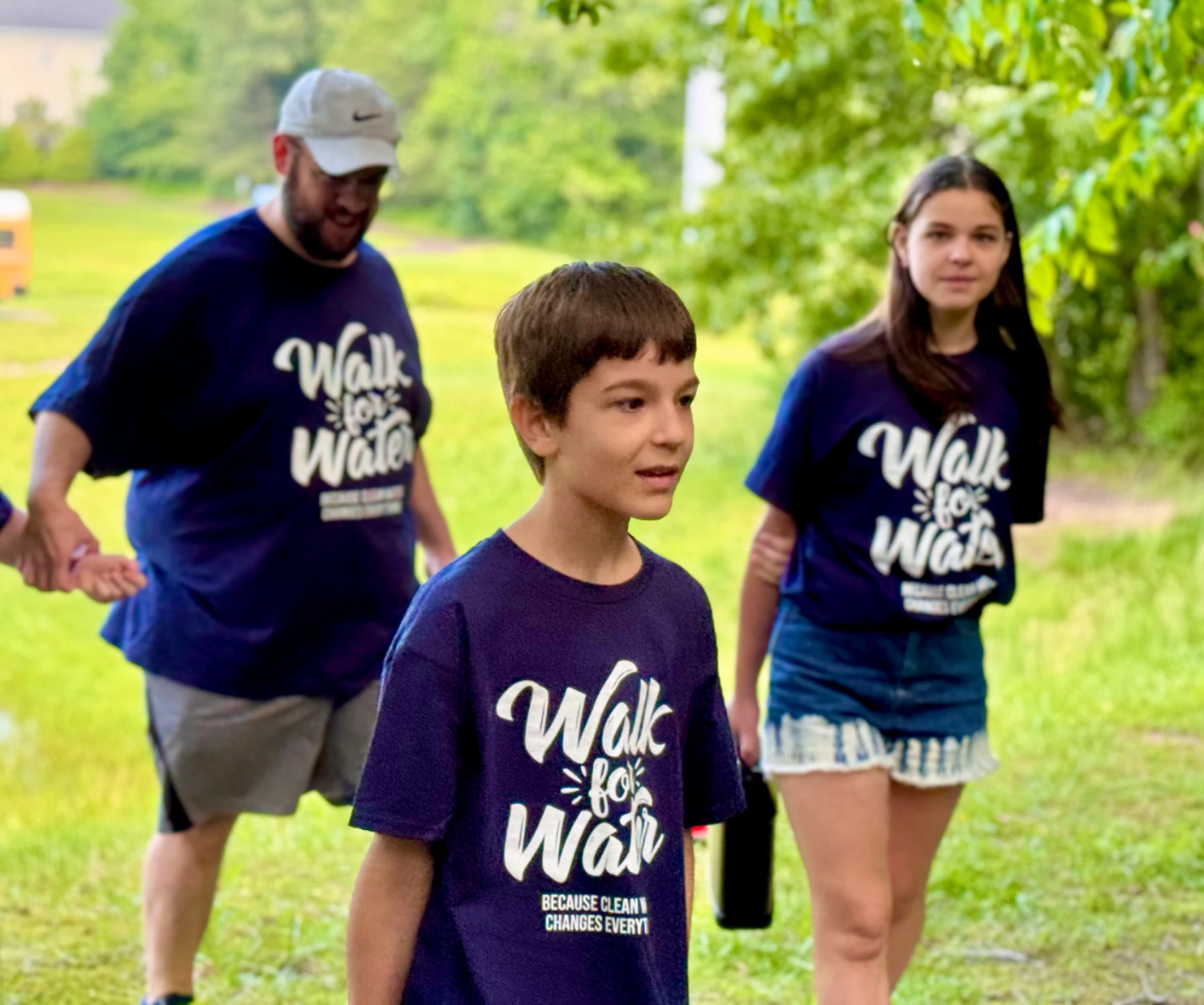 A young boy, a girl, and a man wear matching blue "Walk for Water" t-shirts while walking outdoors in a grassy area for a charity event.