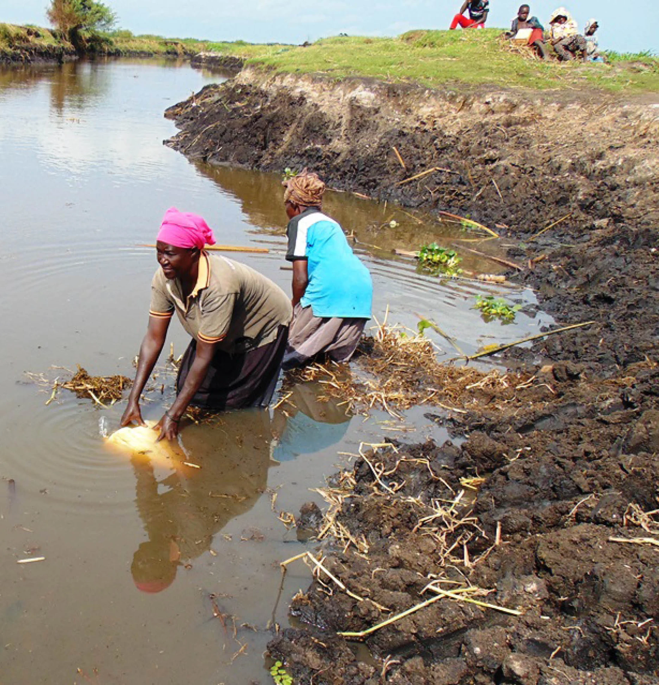 Two African women wade in muddy river water. One fills a yellow plastic jug while others sit on the grassy bank in the background.
