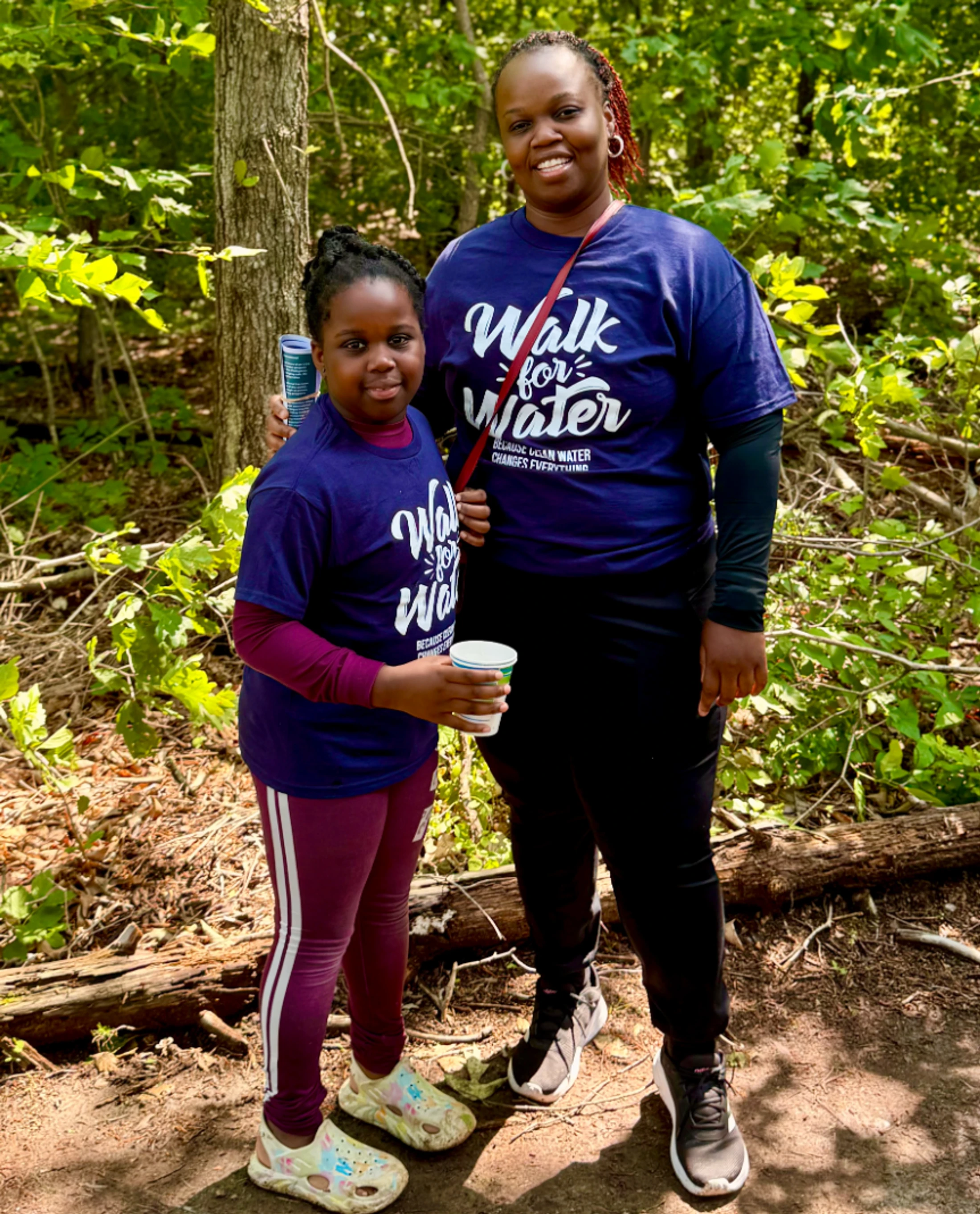 A smiling woman and a young girl in matching 'Walk for Water' t-shirts pose together on a wooded trail during a sunny day.