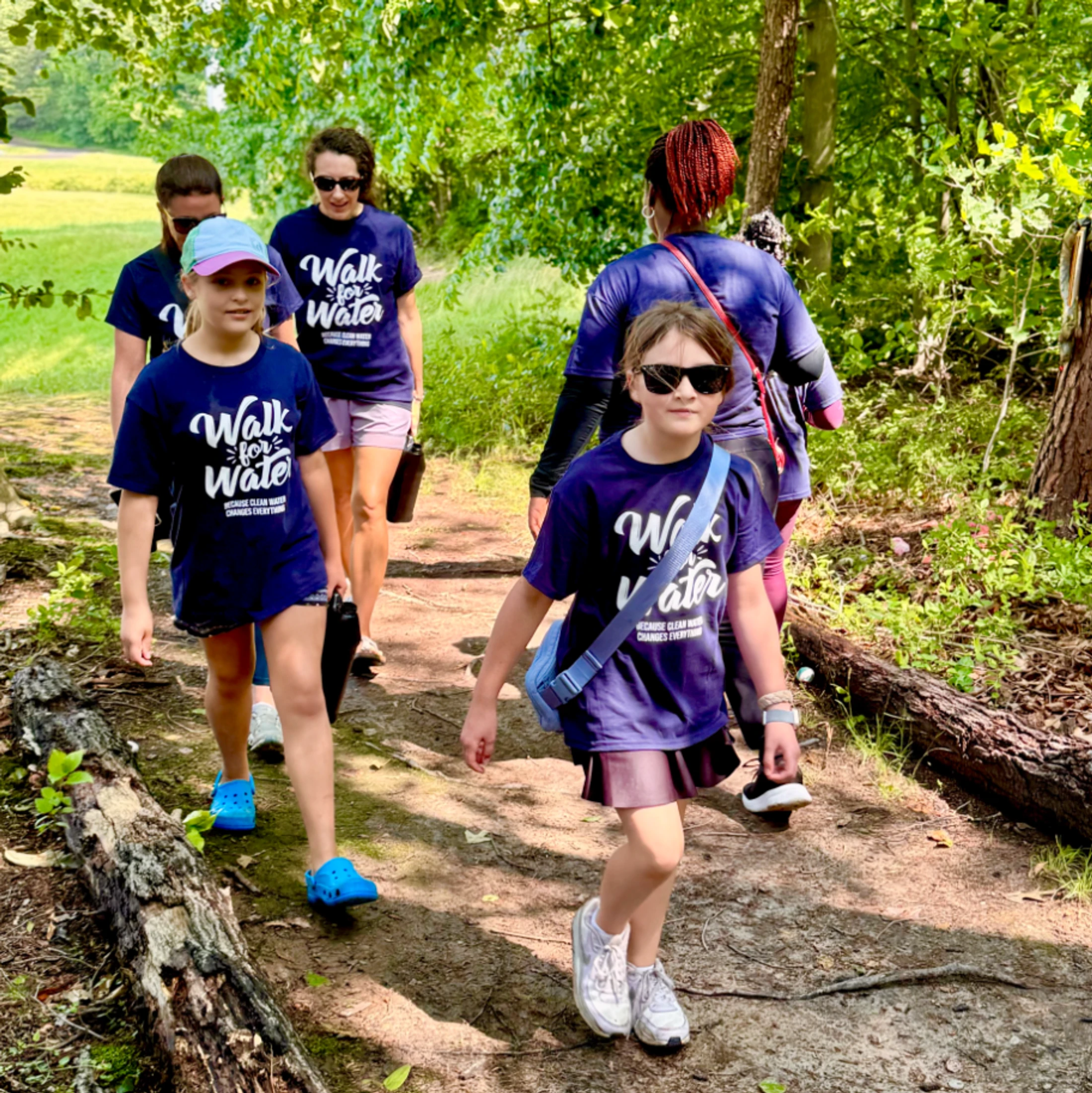 A group of adults and two young girls in matching 'Walk for Water' t-shirts walk along a dirt path in a sunny, wooded area for a charity event.