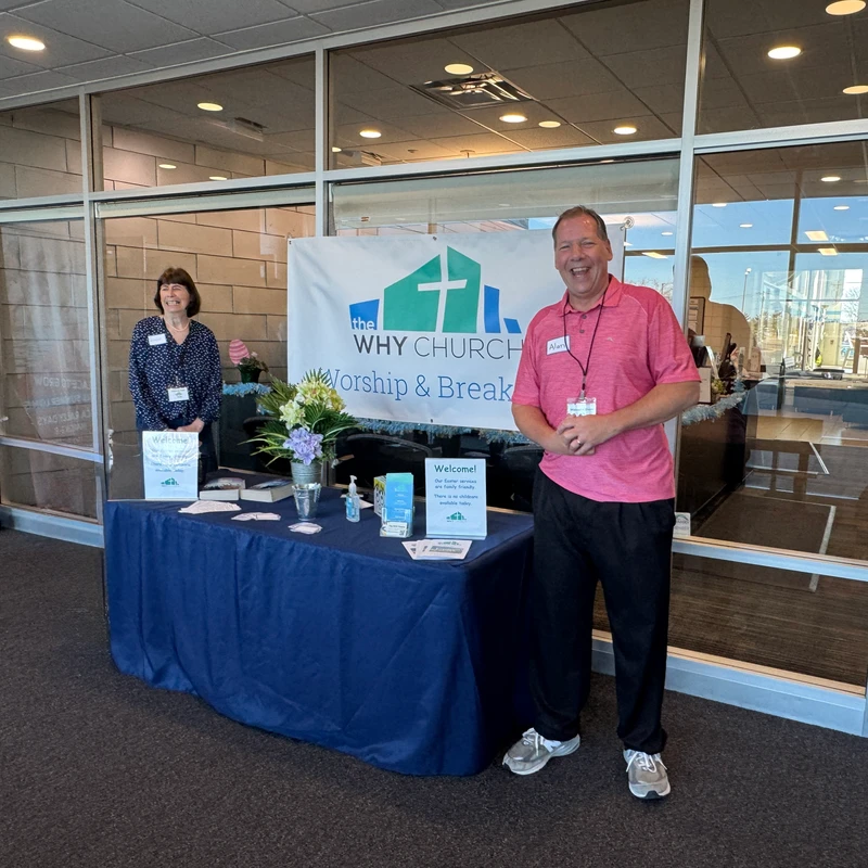 A smiling man and woman stand at a welcome table for 'The WHY Church' in front of a large windowed entrance, ready to greet visitors.