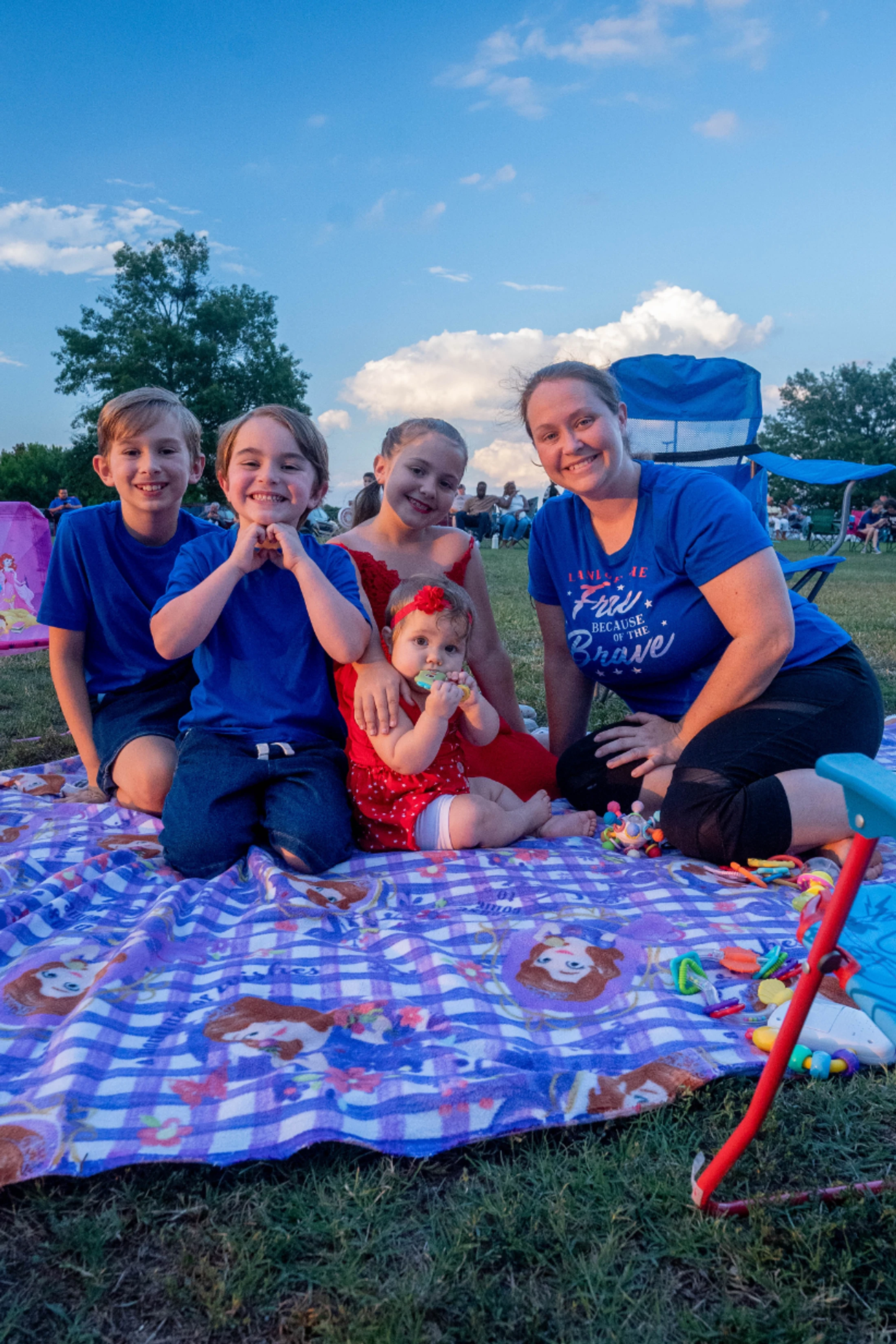 A smiling woman and four children, two boys, a girl, and a baby, sit on a purple blanket on the grass outdoors, posing for a photo under a blue sky.