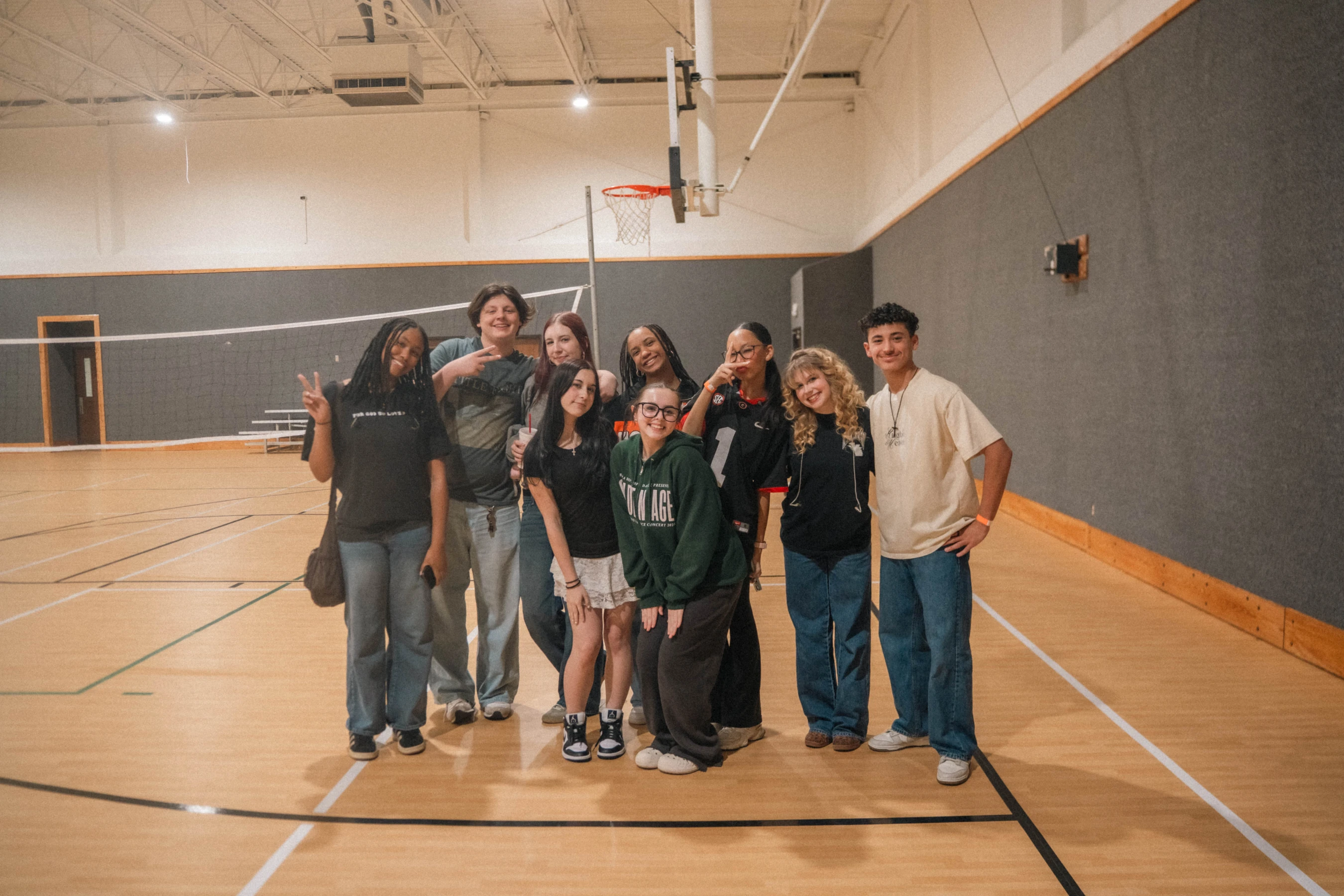A diverse group of nine young people stand together and smile for a photo on a wooden basketball court, with a volleyball net behind them.