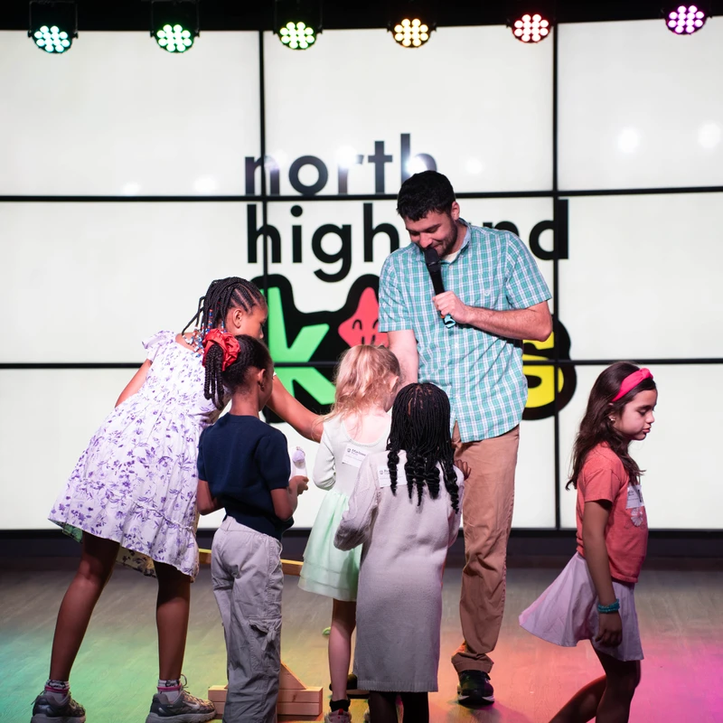 A man in a plaid shirt holds a microphone and smiles, surrounded by a diverse group of young children on a brightly lit stage with a large screen.