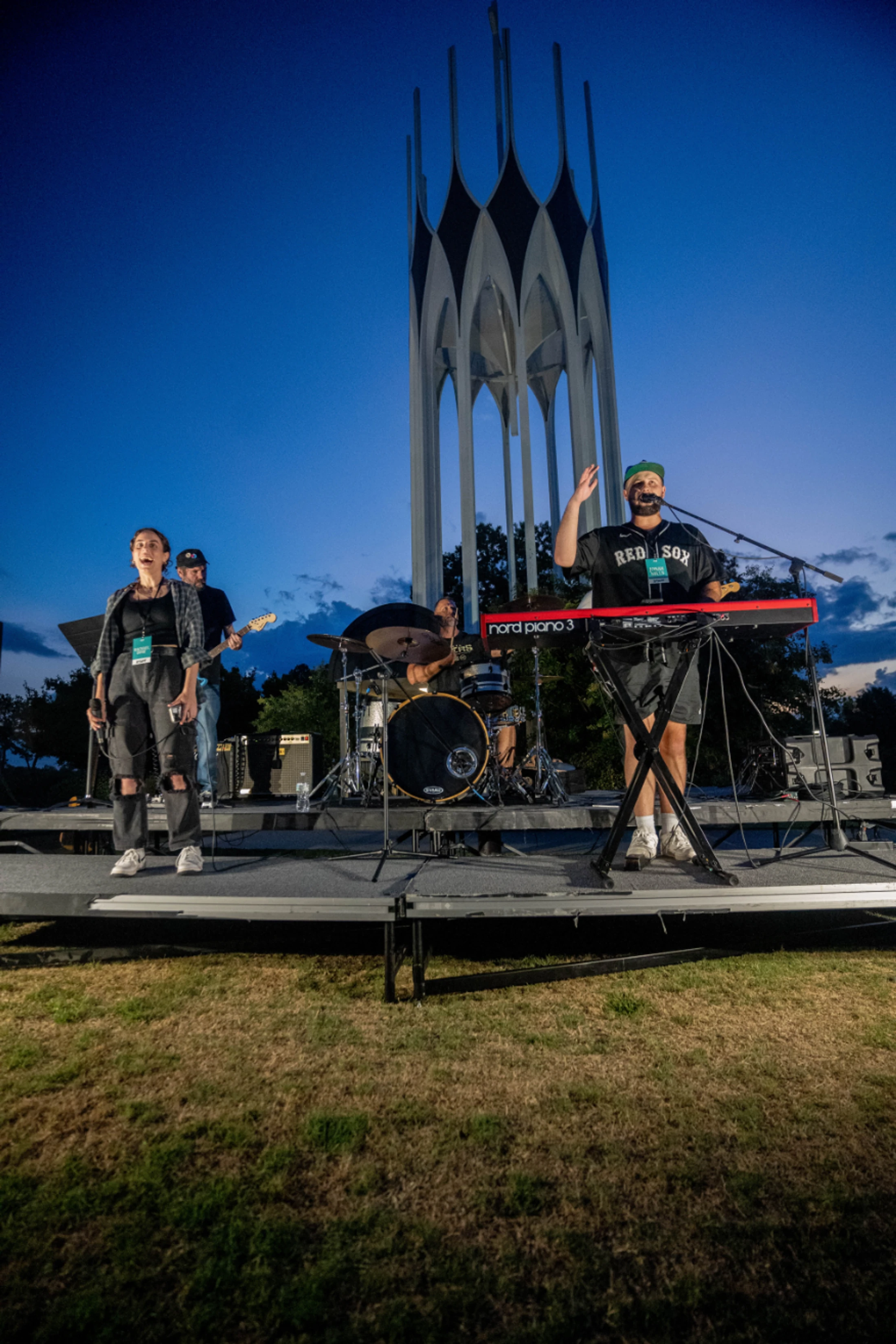 A band with singers, a keyboardist, a drummer, and a guitarist performs on an outdoor stage at dusk in front of a tall, modern white sculpture.