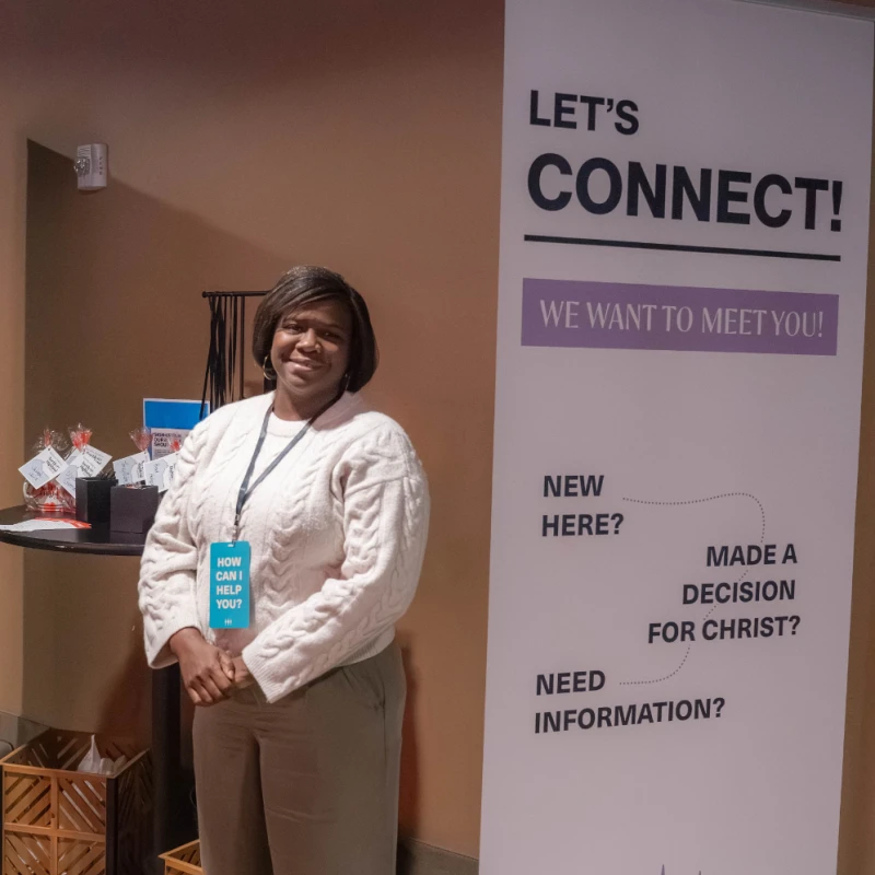 A smiling woman stands next to a large banner that reads 'Let's Connect! We want to meet you!' at a church welcome station.