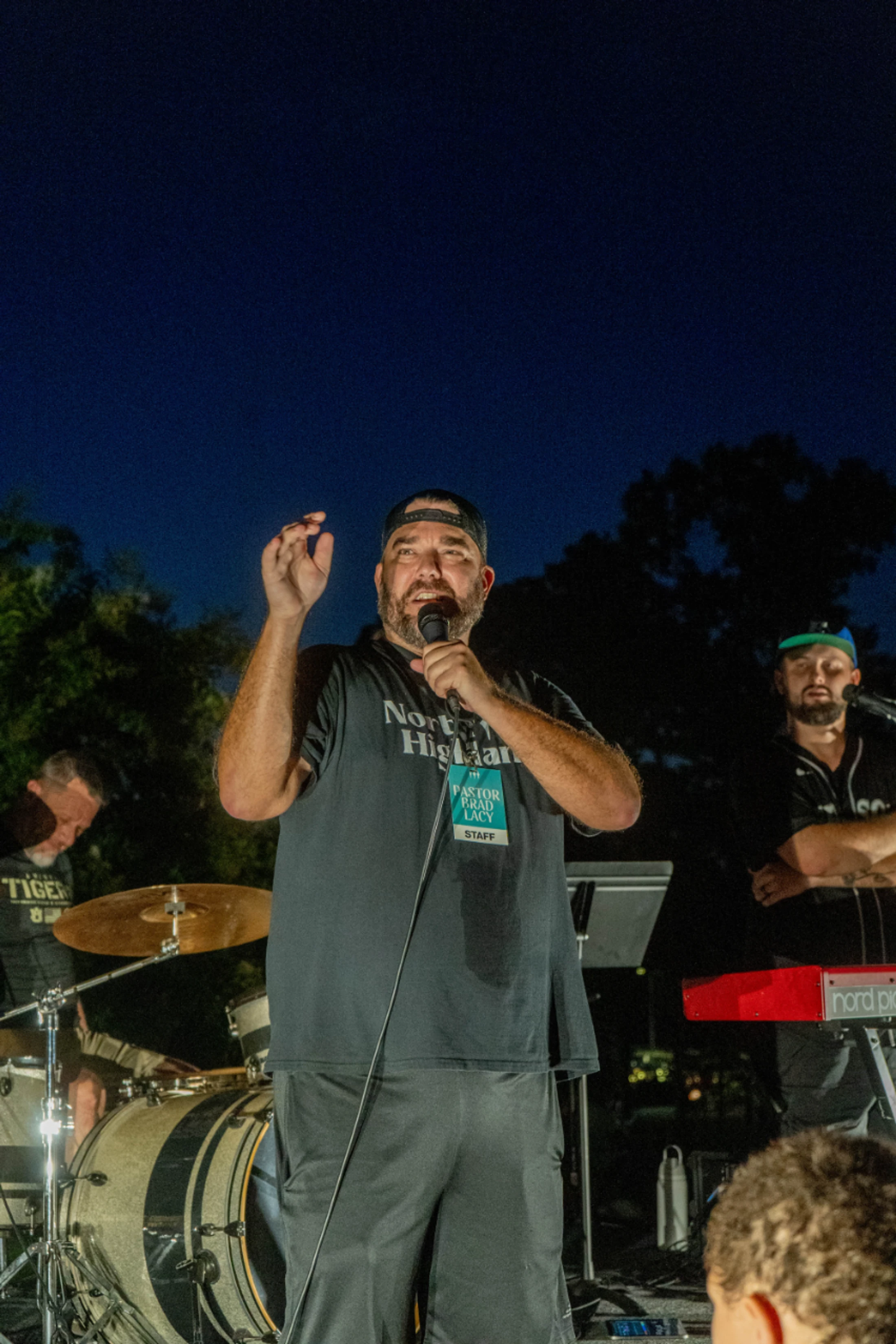 A man in a backward baseball cap speaks into a microphone on an outdoor stage at night, with a drummer and other band members behind him.