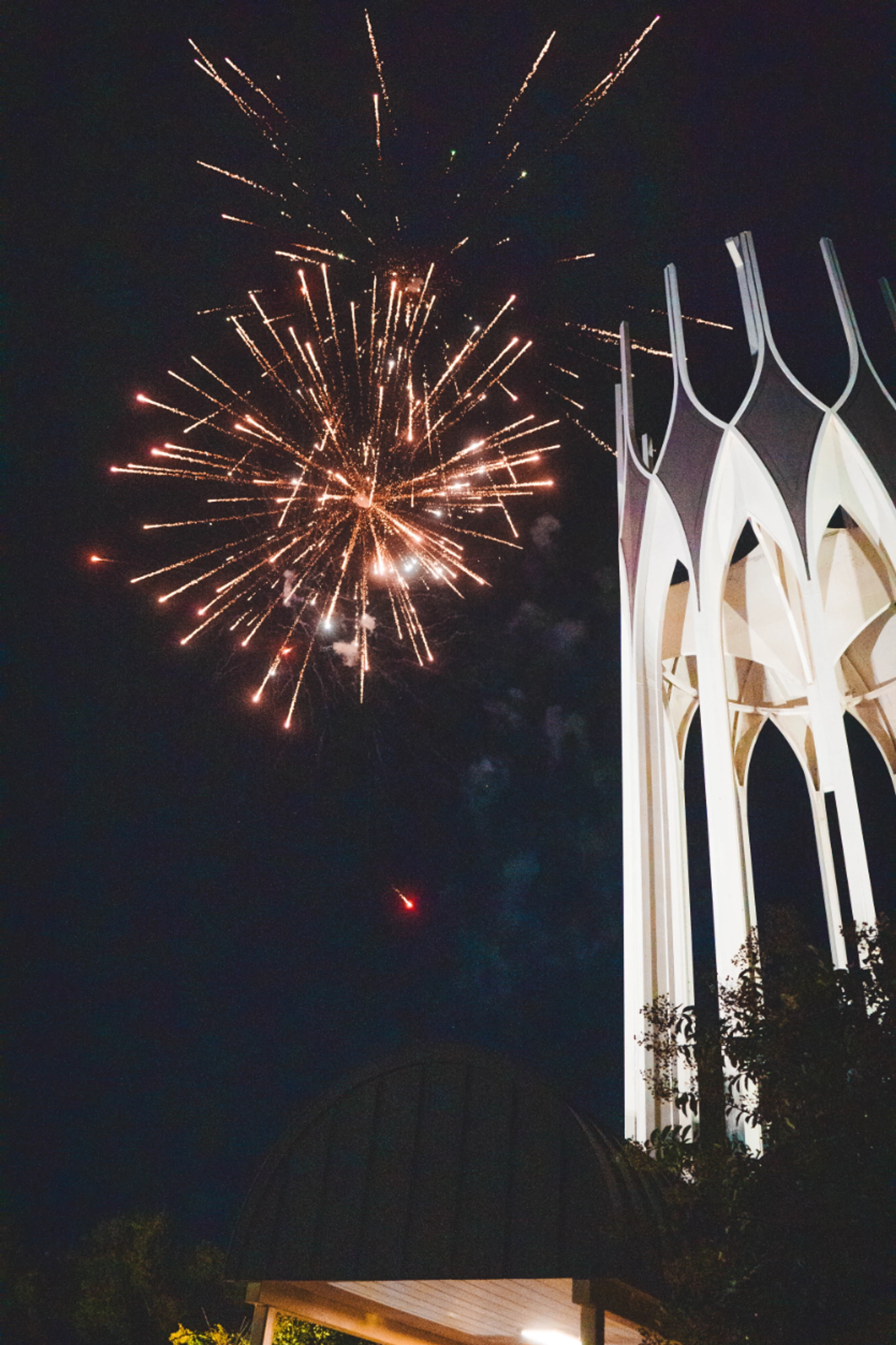 Golden fireworks burst in the dark night sky next to a tall, illuminated white bell tower with modern, pointed arches.