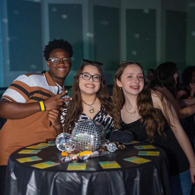 Three diverse teenagers, one boy and two girls, smile for the camera while standing behind a table decorated with small disco balls at a party.