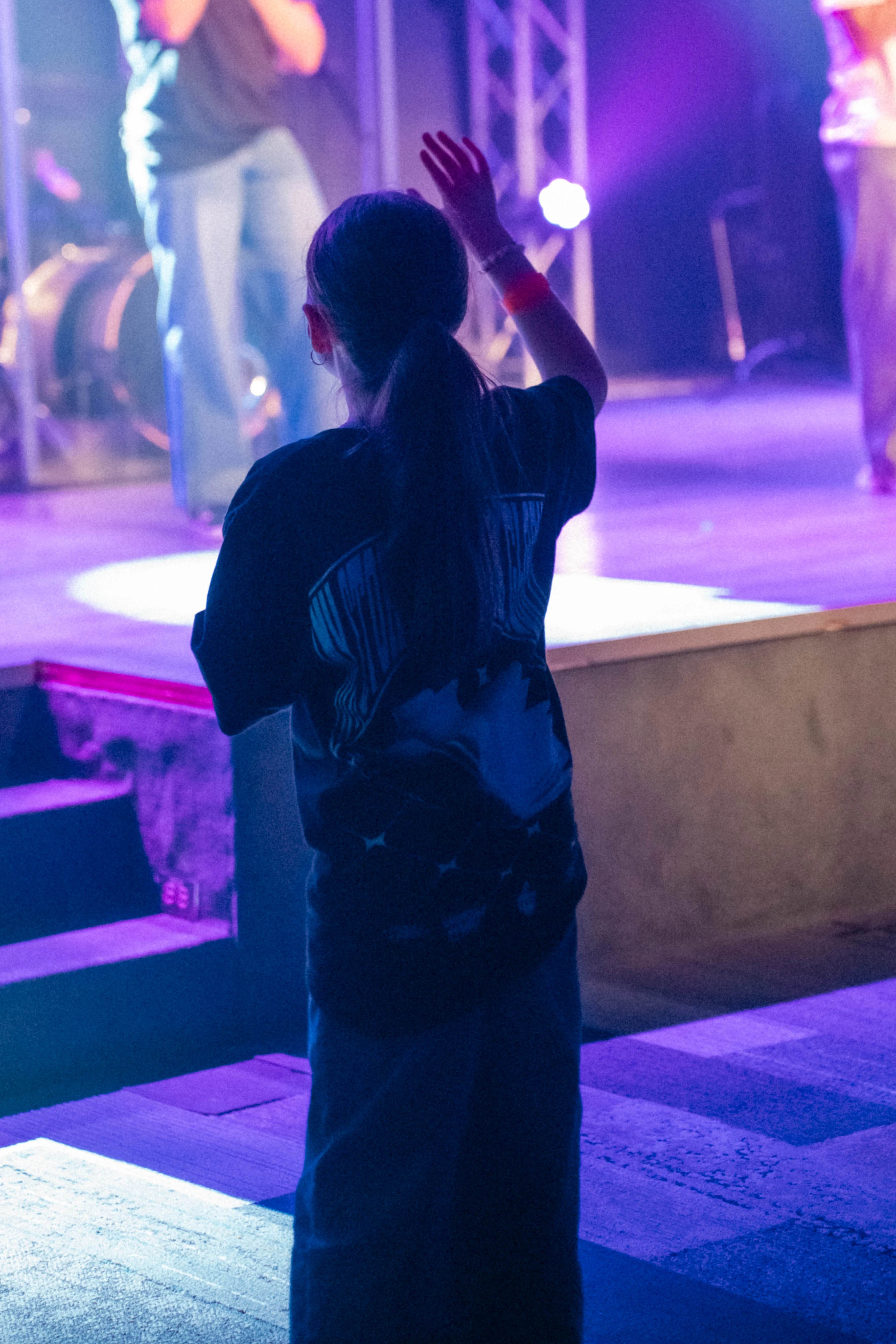 The back of a young person with a ponytail raising a hand in worship while facing a stage bathed in purple and blue light during a performance.
