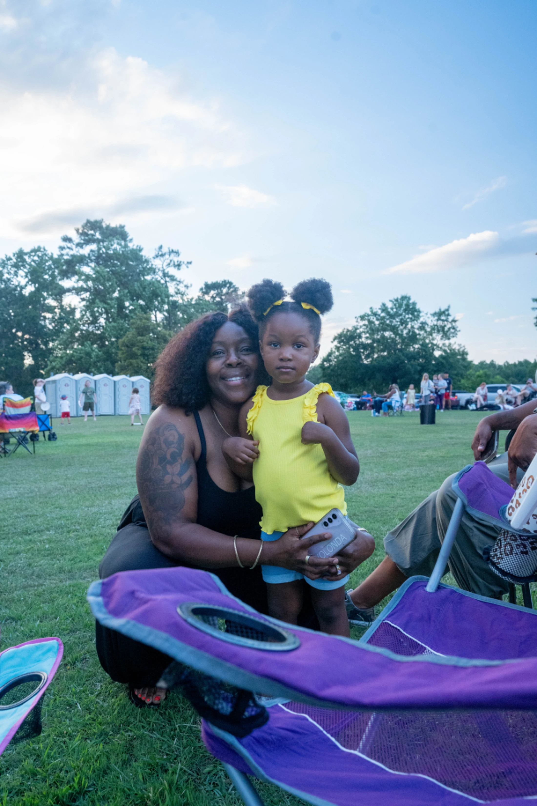 A smiling woman with curly hair and an arm tattoo kneels on the grass and hugs a young girl in a yellow shirt who is holding a phone at a park event.
