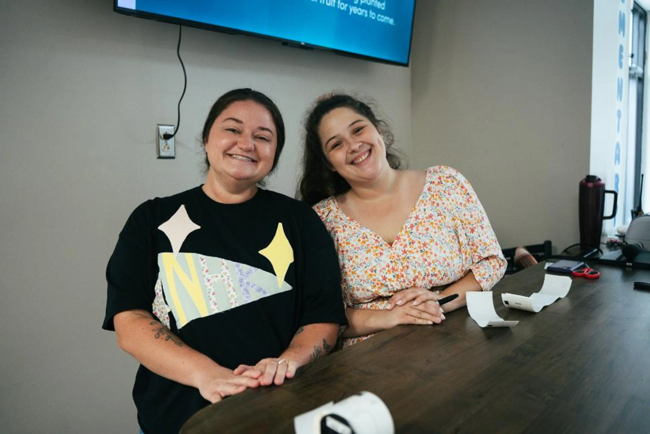 Two women sit side-by-side at a dark wooden table in an office, smiling warmly at the camera. One wears a graphic t-shirt, the other a floral top.