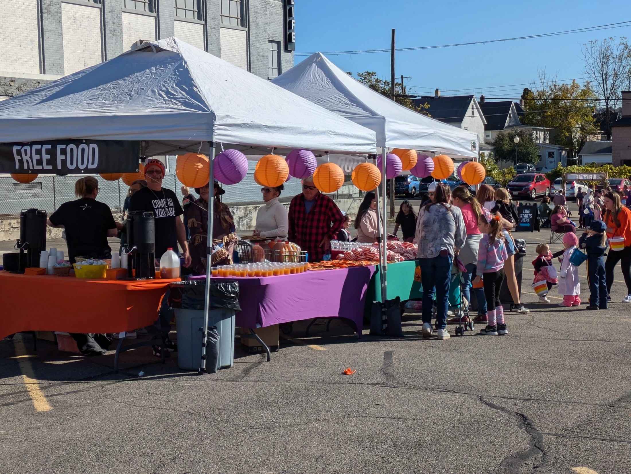 A crowd of people and children in costumes gather at an outdoor community event with tents offering free food and drinks under a clear blue sky.