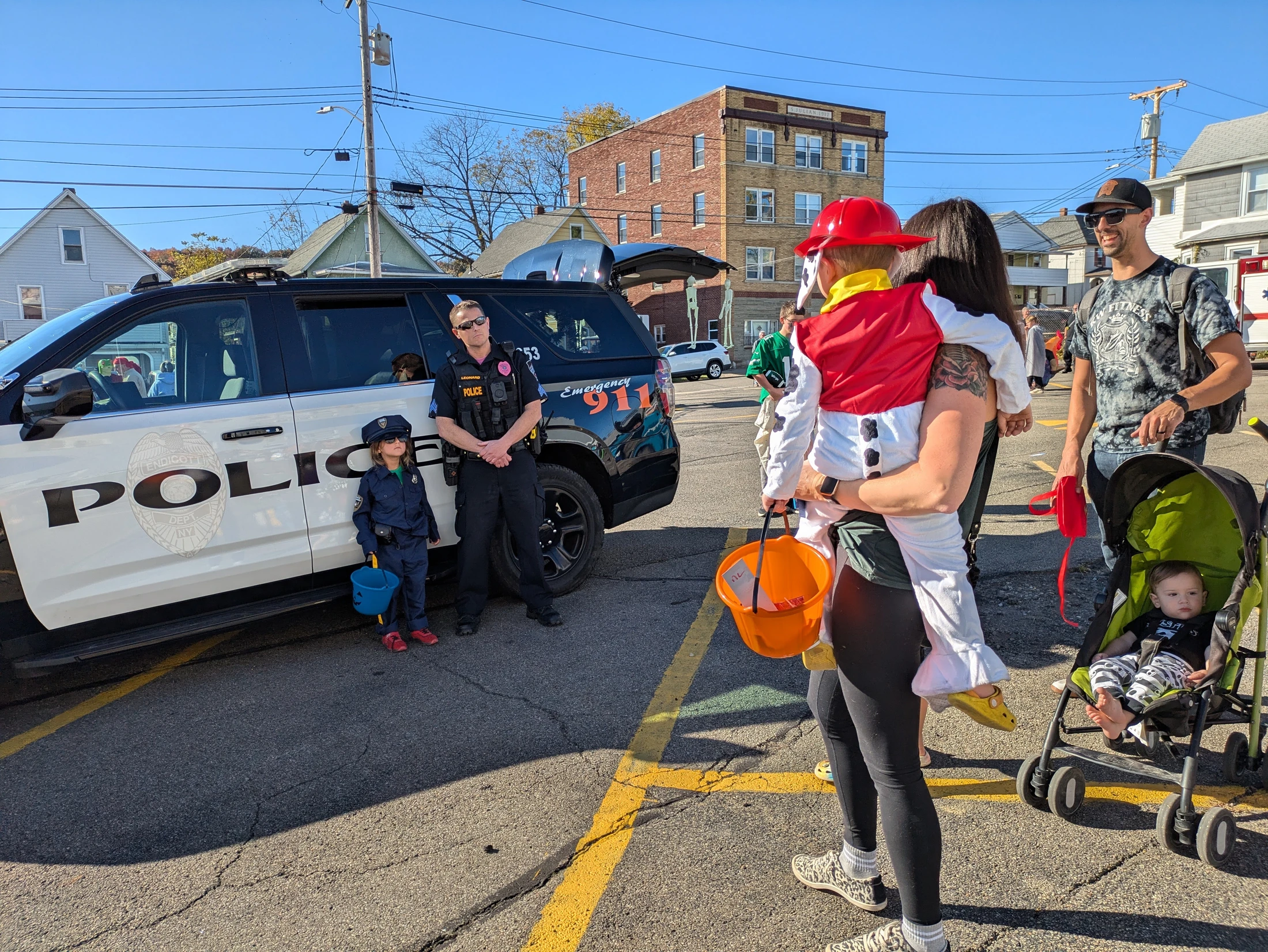 A police officer stands next to his car with a small child dressed as an officer, while a family with a child in a firefighter costume looks on.