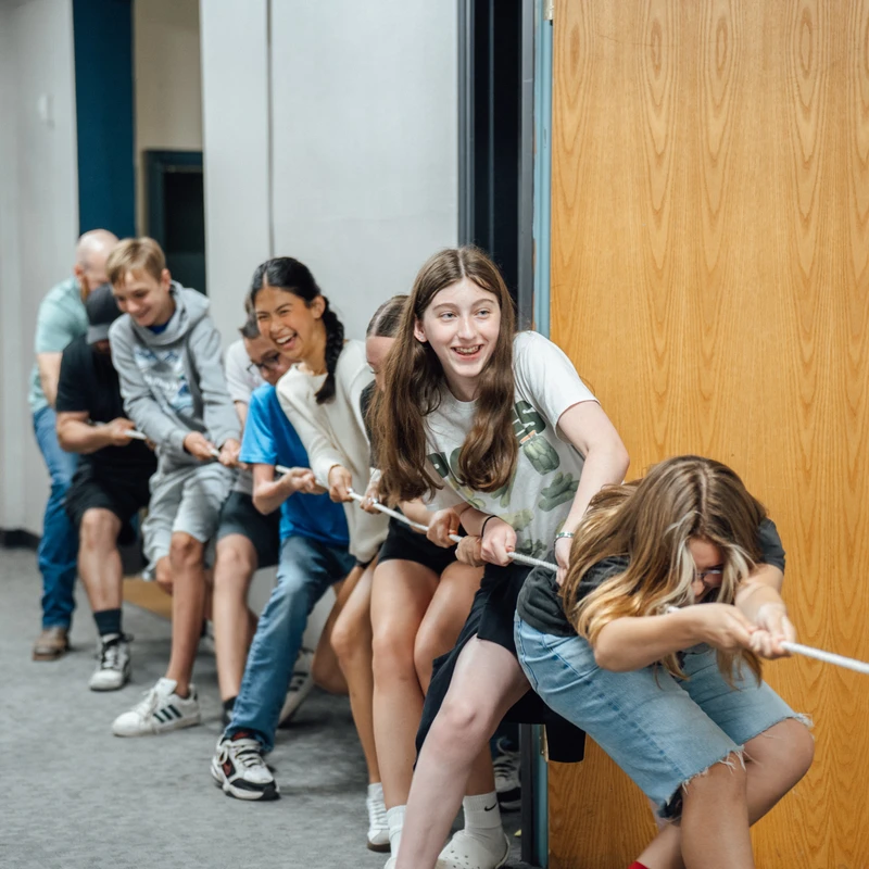 A happy group of young people and adults lined up in a hallway, pulling together on a rope during an energetic game of tug-of-war.