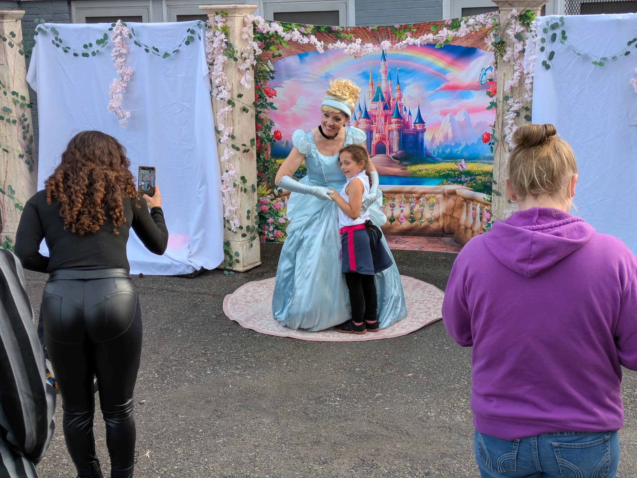 A woman in a Cinderella costume hugs a smiling young girl as they pose for a photo in front of a fairytale castle backdrop at an outdoor event.