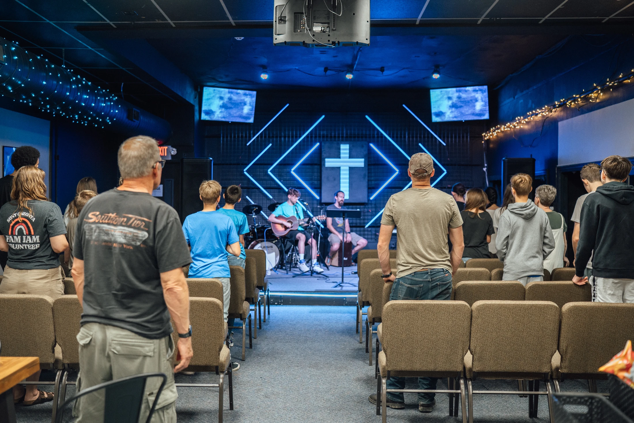View from the back of a modern church where a congregation stands and watches a band perform on a stage lit with blue neon lights and a large cross.