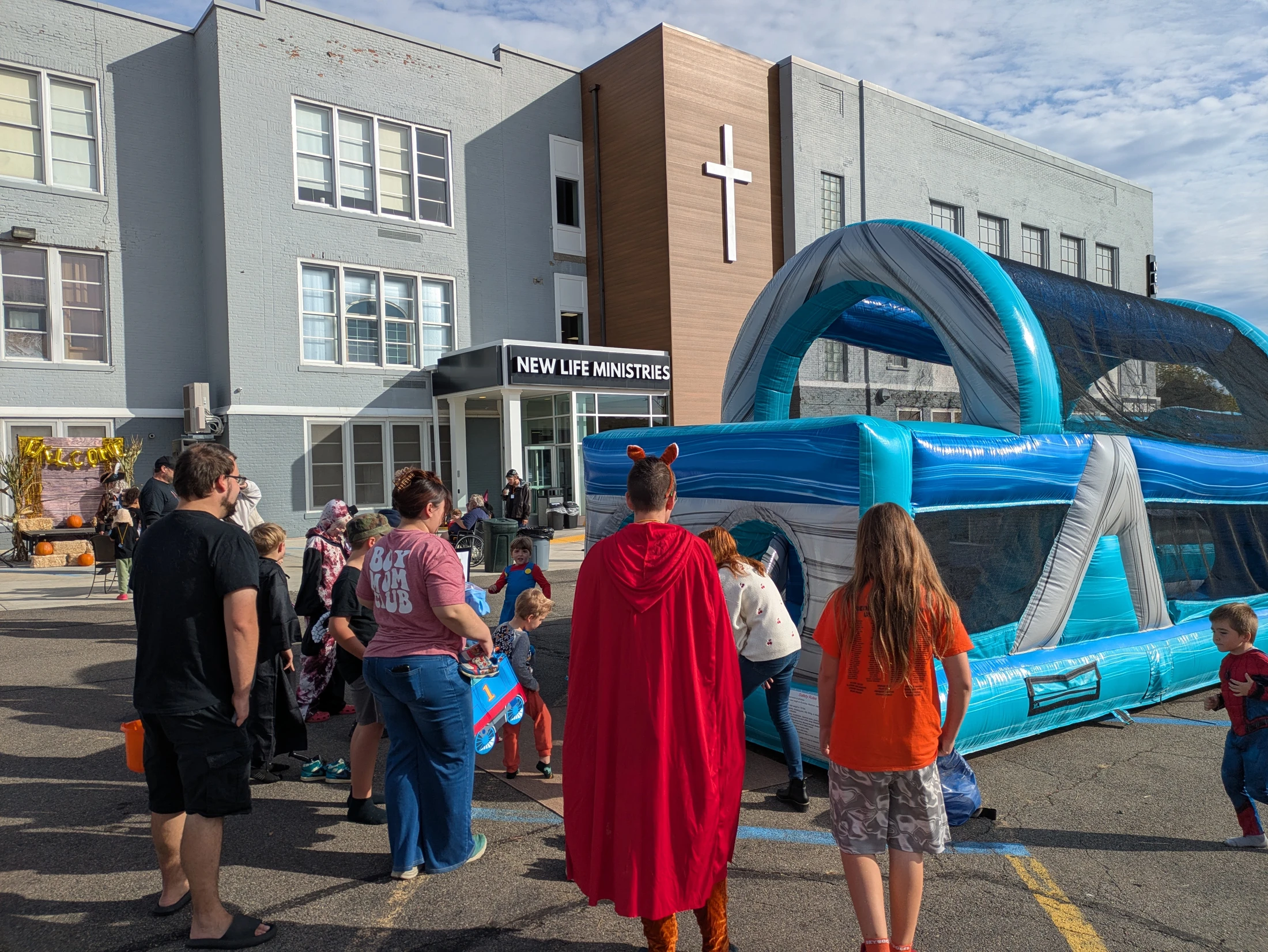 Families and children in costumes gather in a church parking lot for a fall festival, with a large blue and grey inflatable bounce house in the foreground.