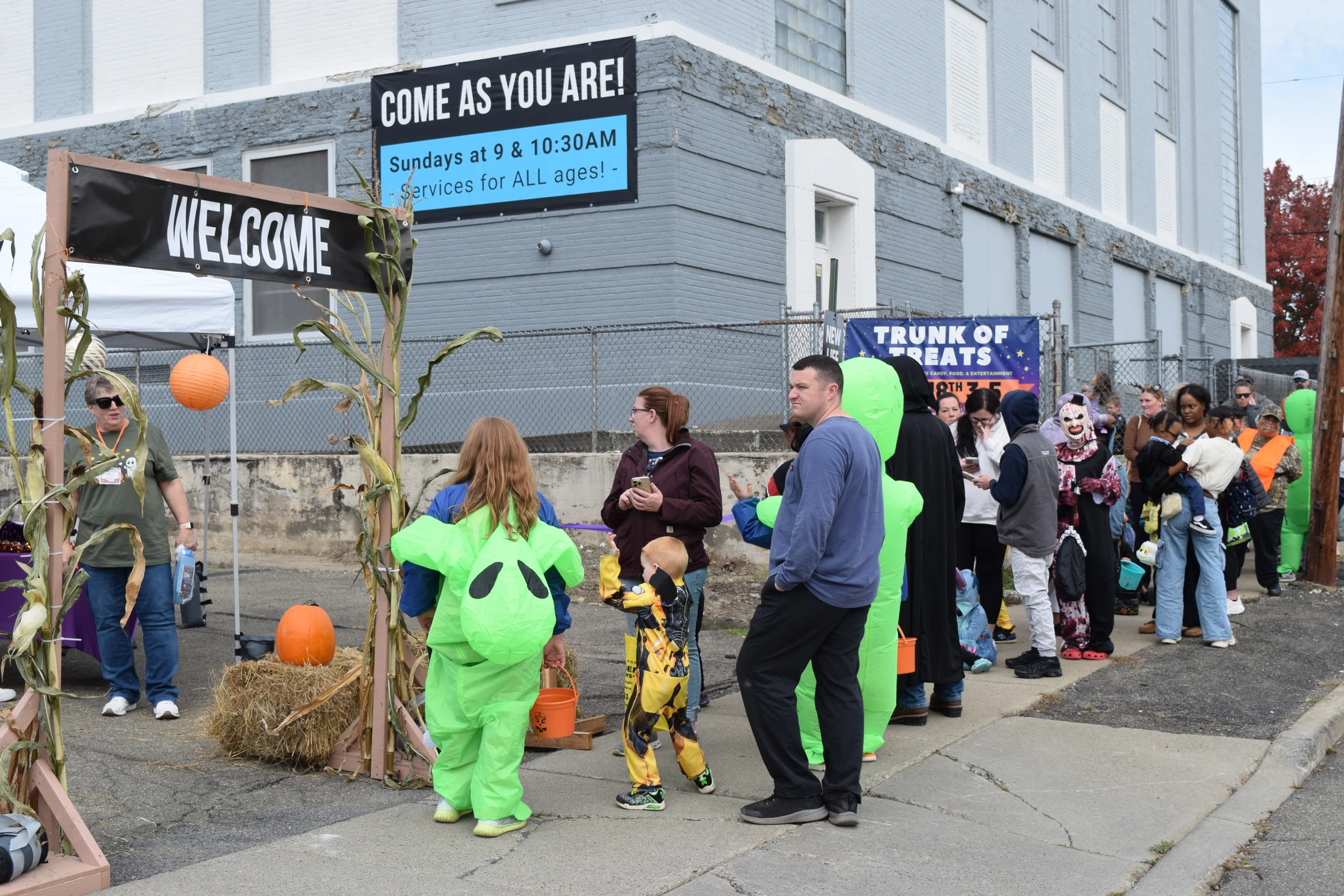 A diverse group of adults and children in Halloween costumes wait in line on a sidewalk for a trunk or treat event outside a large brick building.