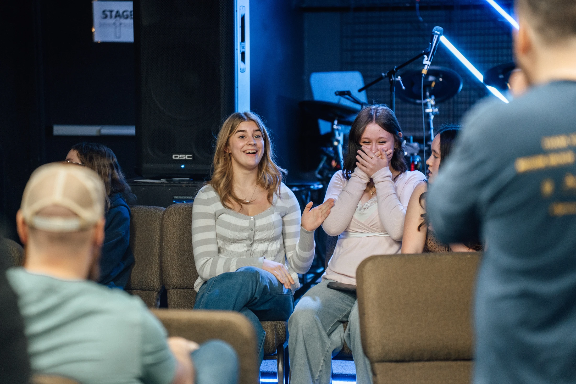 Two young women sit in an audience and laugh. One has a wide smile while the other covers her mouth with her hands. A stage with blue lights is behind them.
