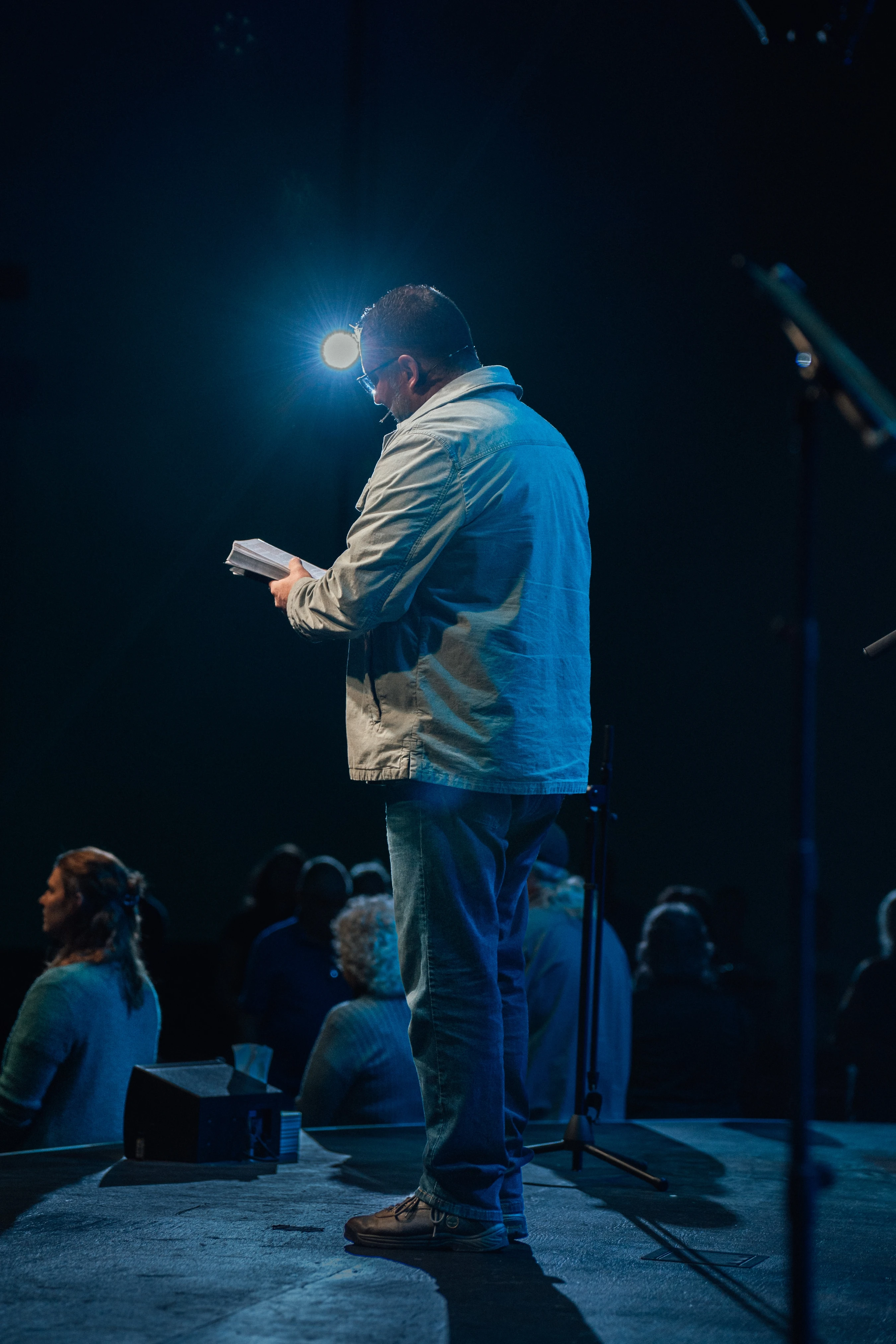 A man in a jacket and jeans stands on a dark stage, reading from papers in his hands. He is illuminated from behind by a single bright, blue spotlight.