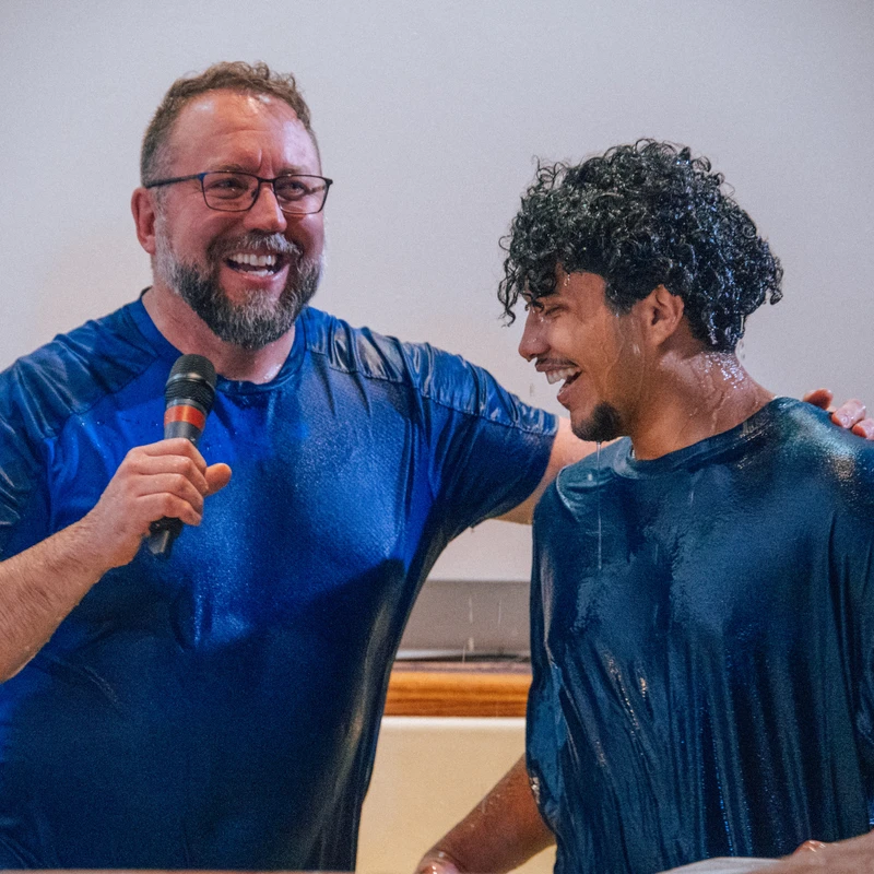 A smiling pastor with a microphone stands next to a smiling young man in a baptismal font. Both are wearing wet blue shirts during the ceremony.