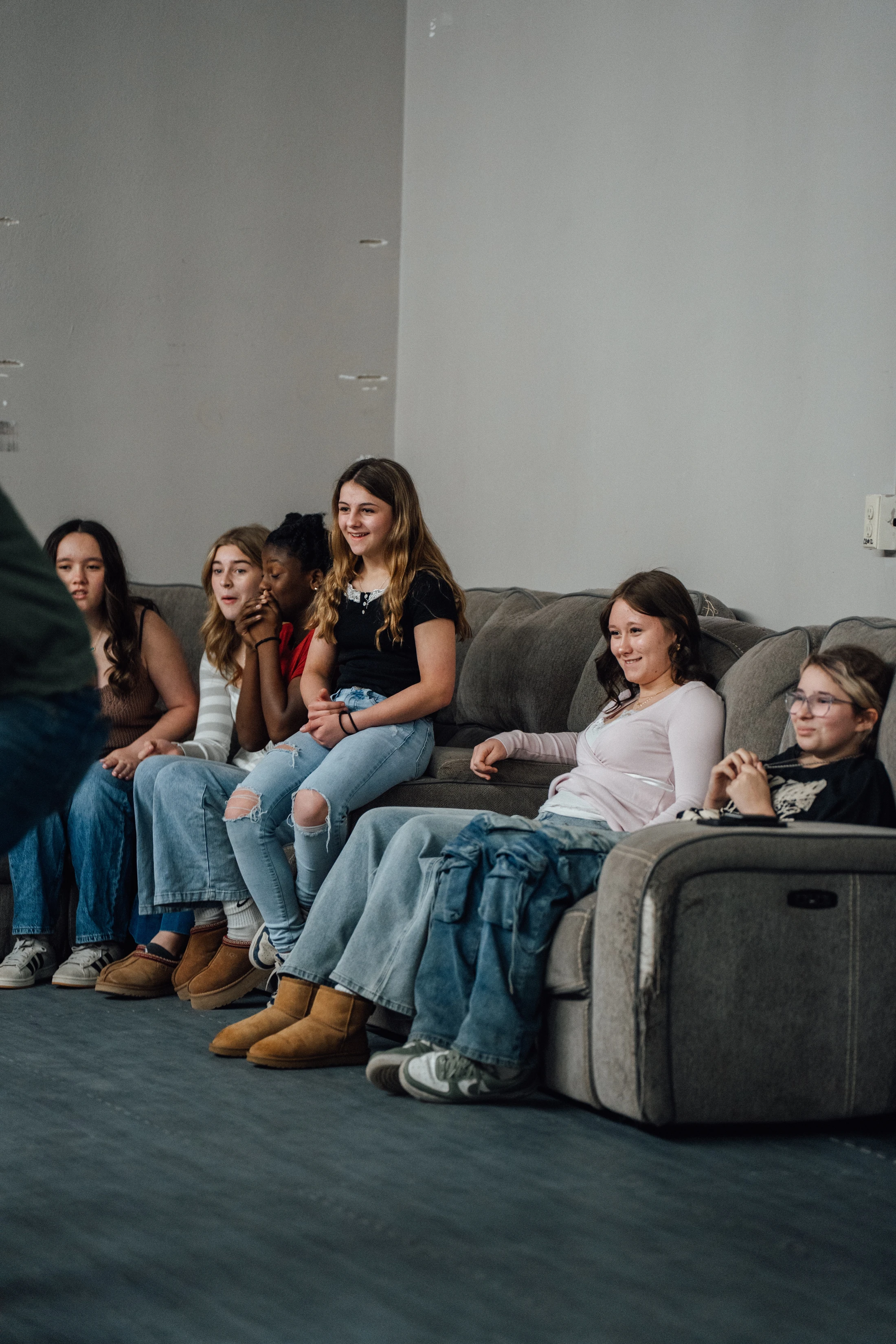 A diverse group of six teenage girls sits closely together on a grey sofa, listening attentively to someone who is out of the frame.