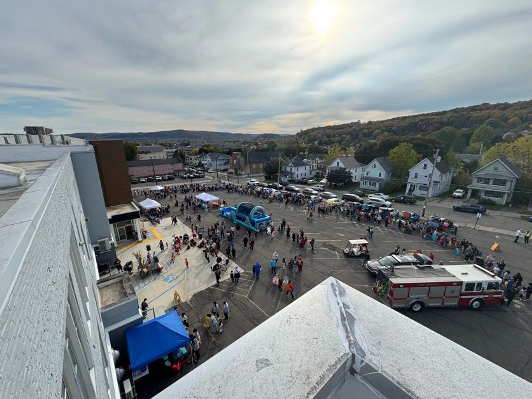 An aerial shot of a bustling town fair in a parking lot, with families, a fire truck, tents, and an inflatable structure under a partly cloudy sky.