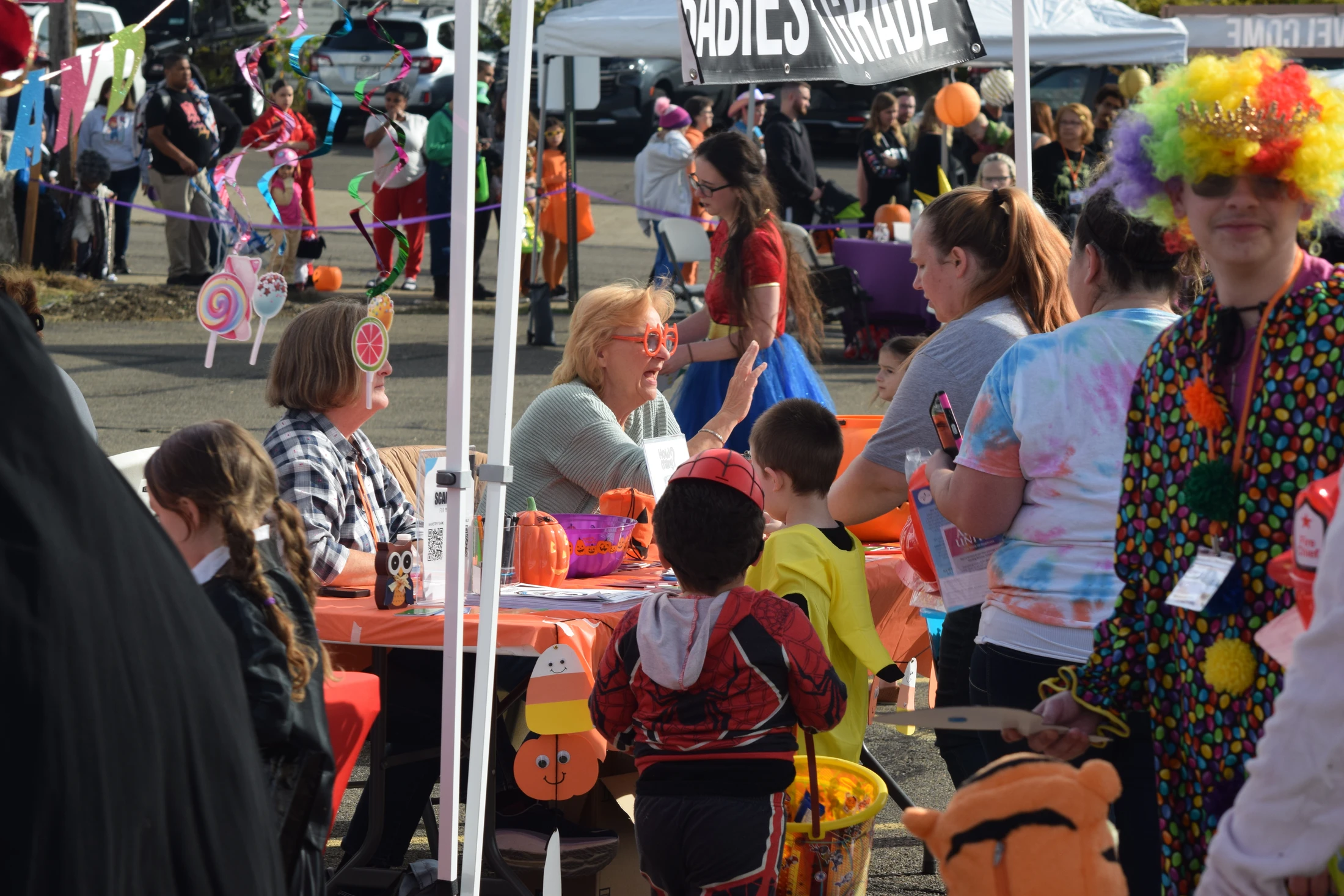 A crowded outdoor Halloween event where children in costumes gather around a table under a tent, interacting with adults. One person wears a rainbow clown wig.