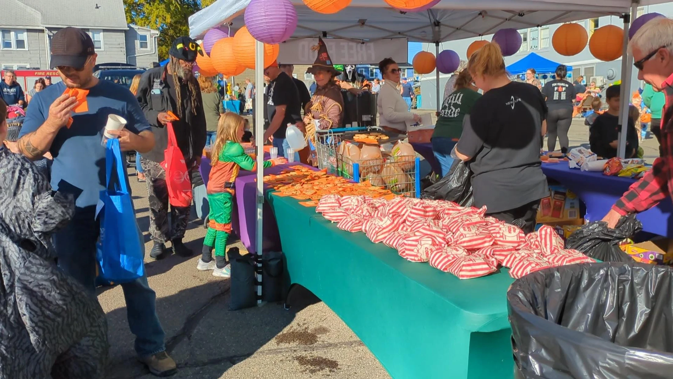 A crowd of adults and children in costumes gather under a tent at an outdoor fall festival, getting free popcorn, cookies, and drinks from a long table.